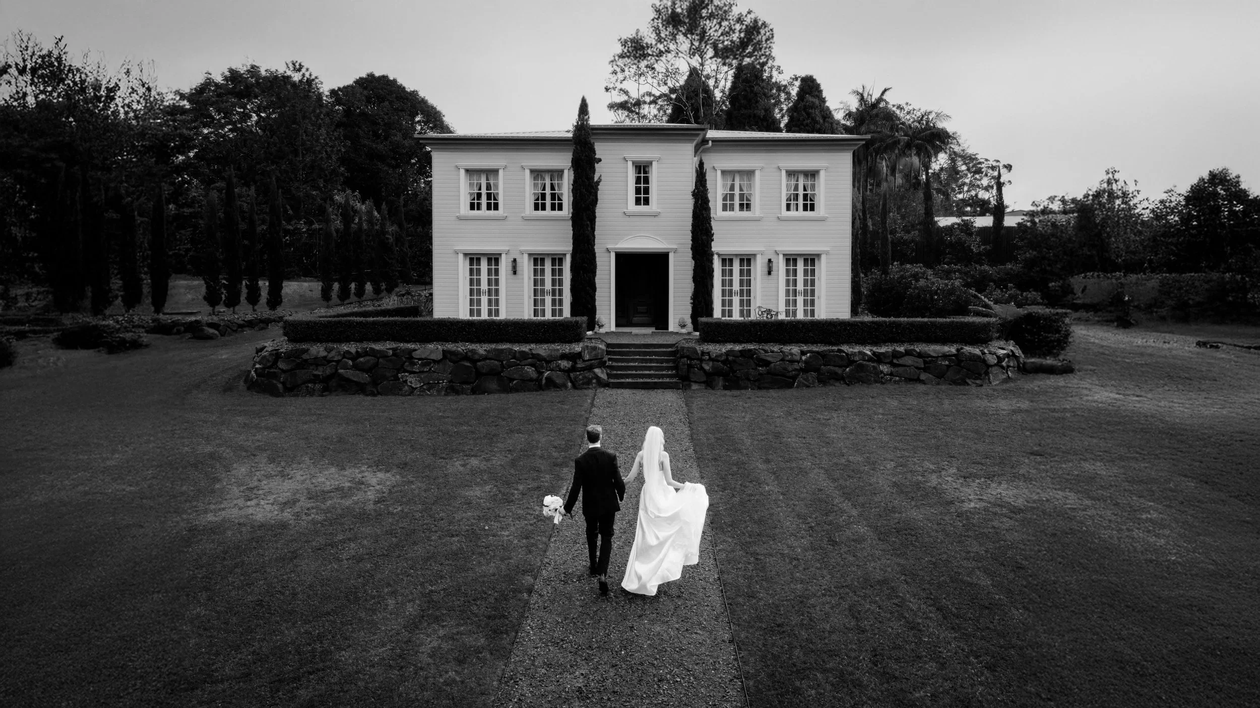 Black and white photo of a bride and groom walking away from a large white house with multiple windows. The bride wears a long dress with a veil, and the groom is in a suit. They hold hands, facing the house with stairs leading up to the entrance, surrounded by a well-manicured lawn and trees.