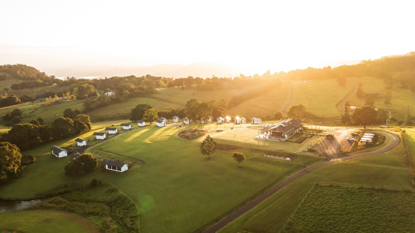 Aerial view of a rural farm with white buildings, a main house, and surrounding green fields at sunset.