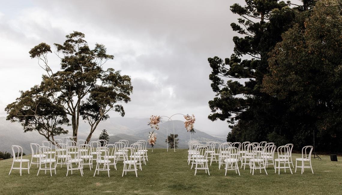Outdoor wedding setup with white chairs arranged on grass, floral arch in the background, and trees on either side with cloudy sky overhead.