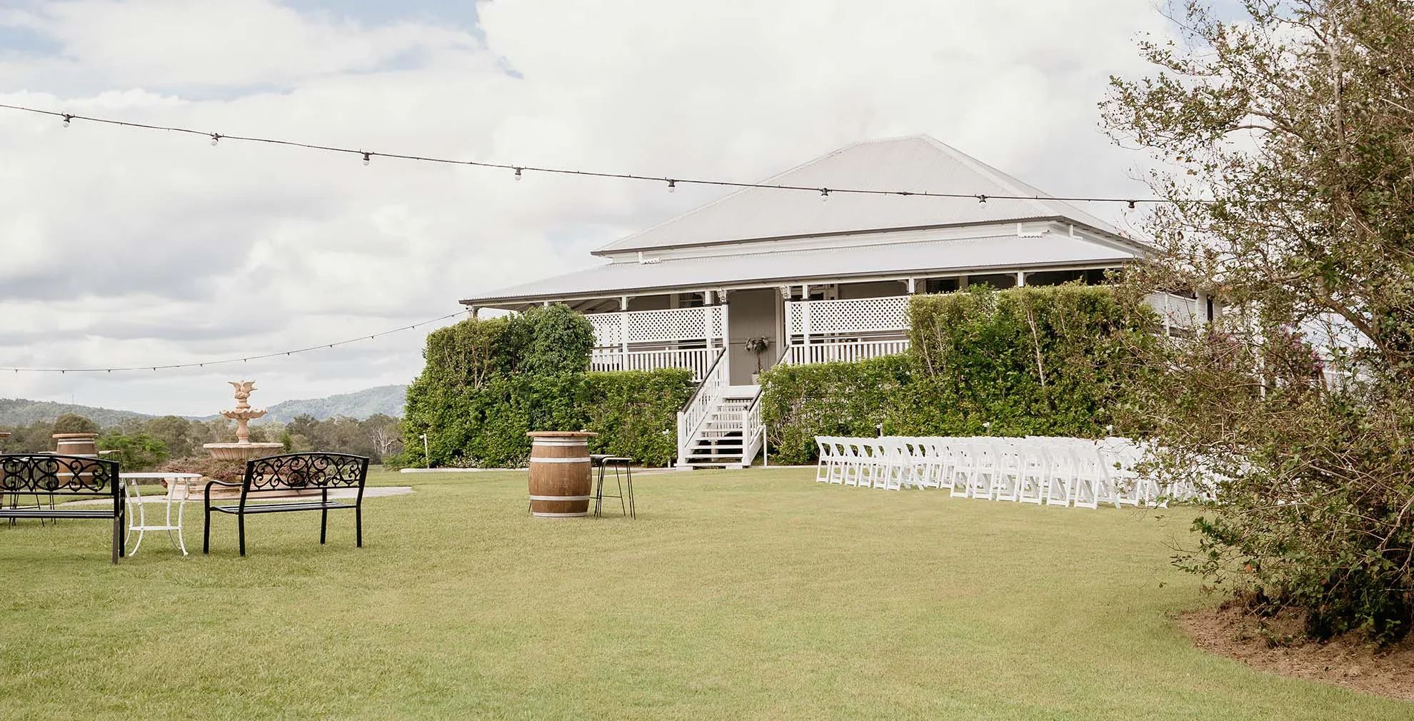 A large white house with a wraparound porch, surrounded by greenery, set on a grassy lawn with outdoor seating and string lights overhead.
