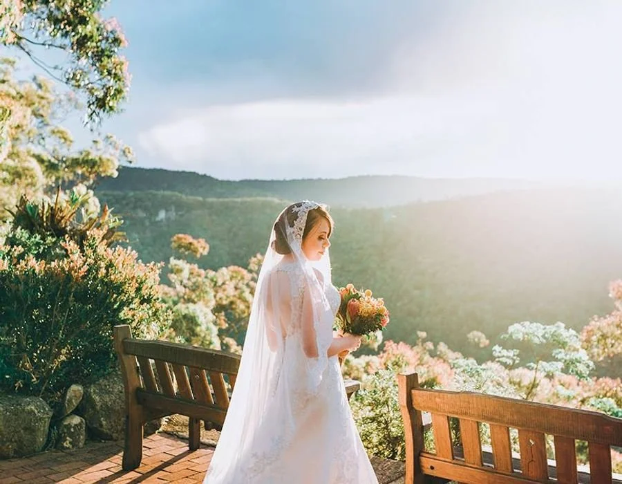 A bride in a white wedding dress and veil holding a bouquet, standing outdoors on a brick pathway near benches with a scenic mountain and flowering bushes background at sunset.