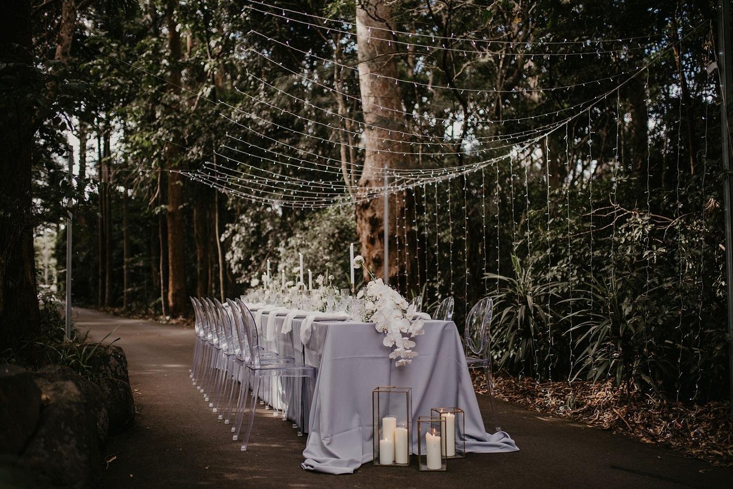 Outdoor wedding reception setup with a long white-draped table decorated with white flowers and candles in lanterns, surrounded by transparent chairs, and hanging string lights overhead, set in a forested area.