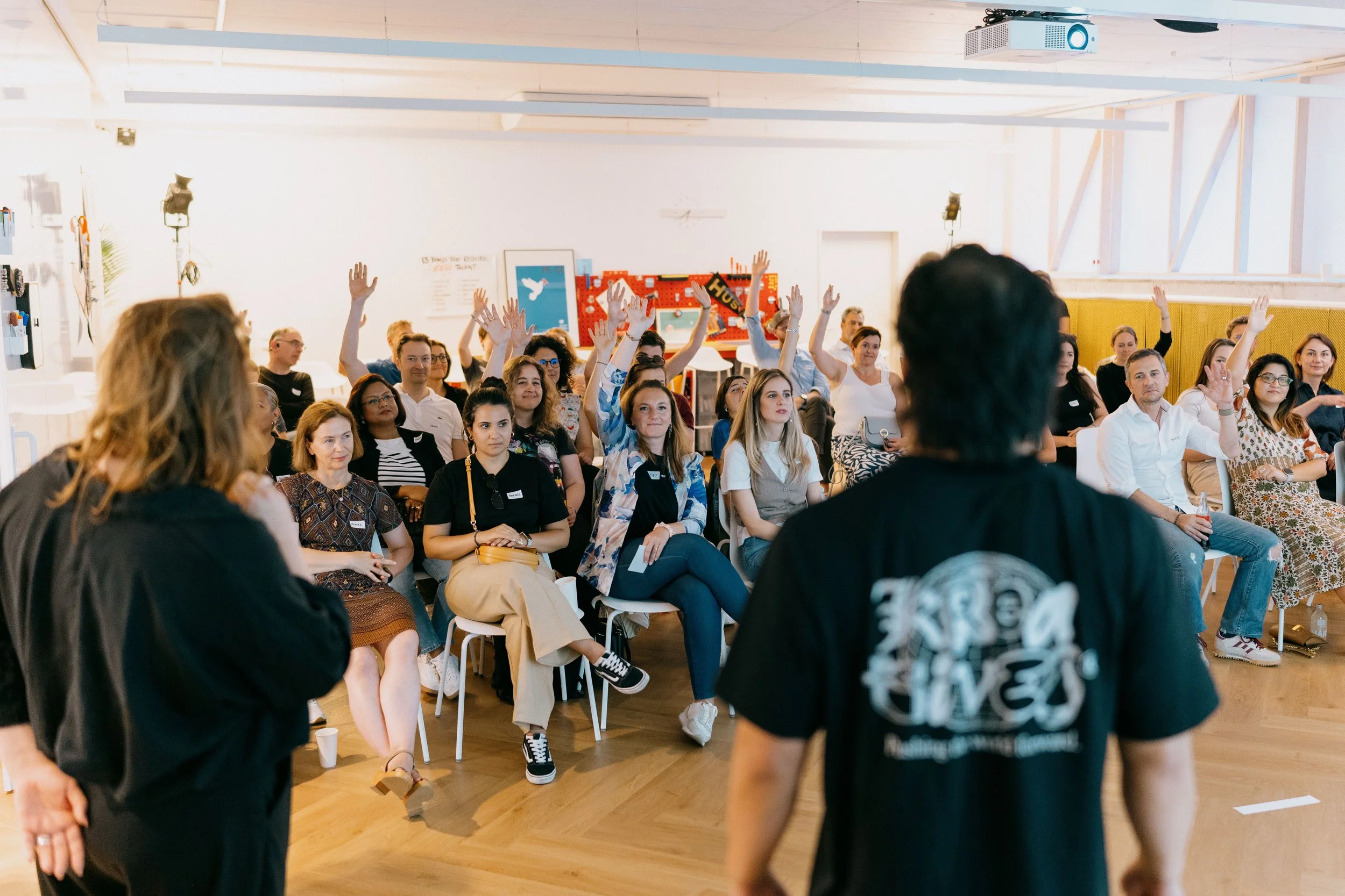 A group of people at a seminar or workshop, seated in chairs facing a speaker, with some participants raising their hands.