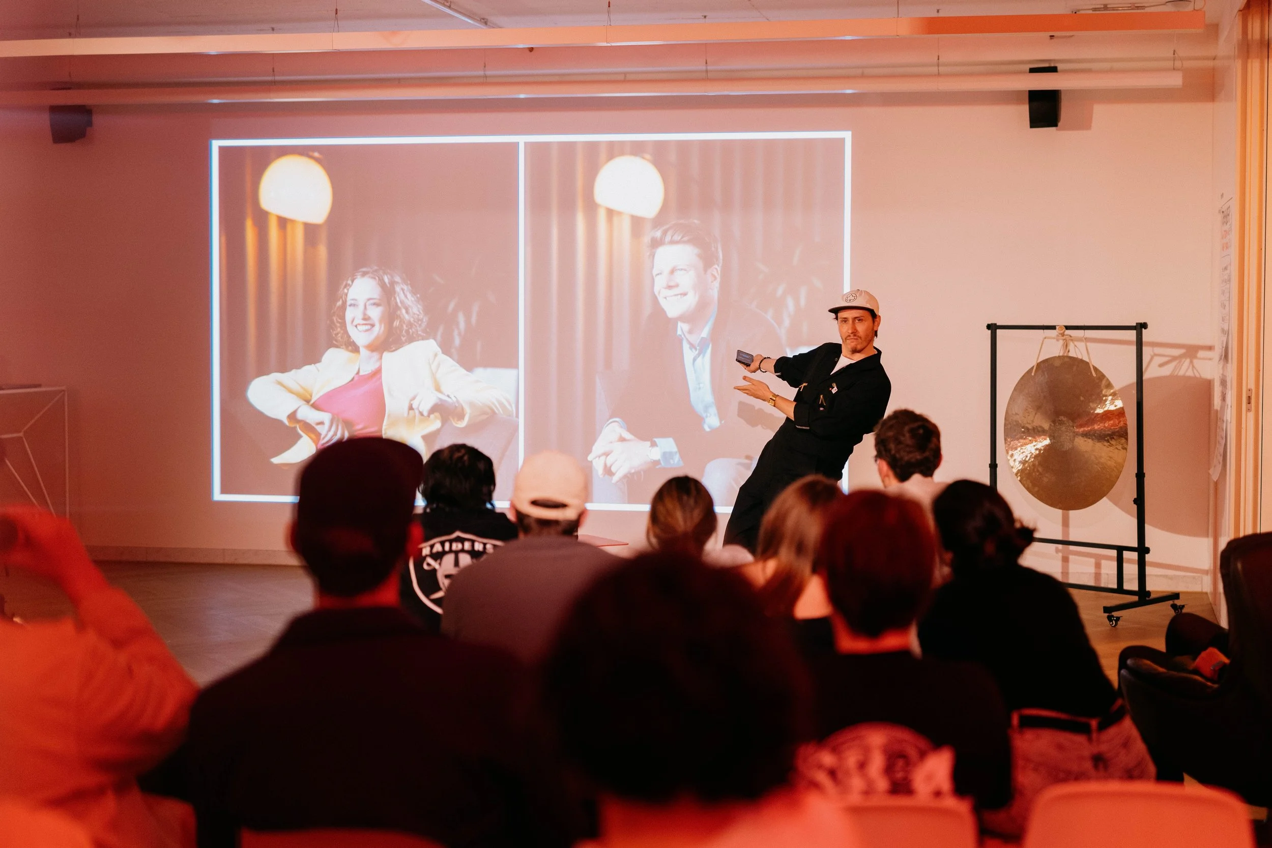 A man giving a presentation to an audience with two large images of people projected on the screen behind him. The images on the screen show a woman with curly hair and a man with a smile, both in a casual indoor setting. The audience is seated in a 