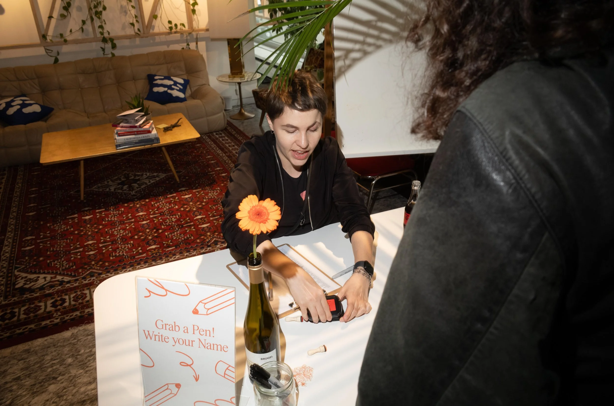A woman sitting at a white table, writing with a pen, with a man standing in front of her. There is a sign on the table that says 'Grab a Pen! Write your Name.' The table has a vase with an orange flower, a bottle, a jar with pens, and a clipboard. T