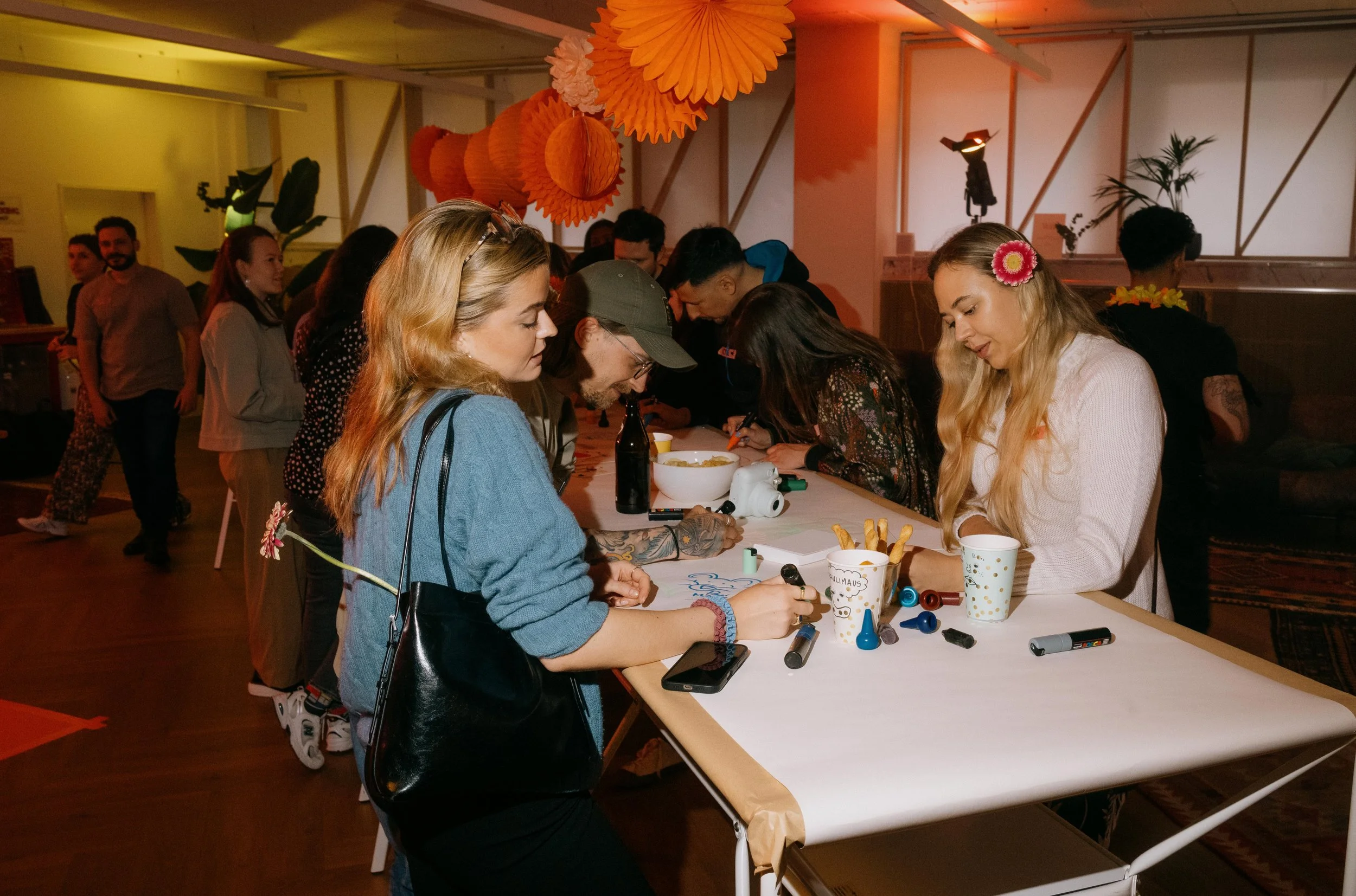 Group of young adults gathered around a table decorating cookies at a party with orange hanging decorations, drinks, and markers.