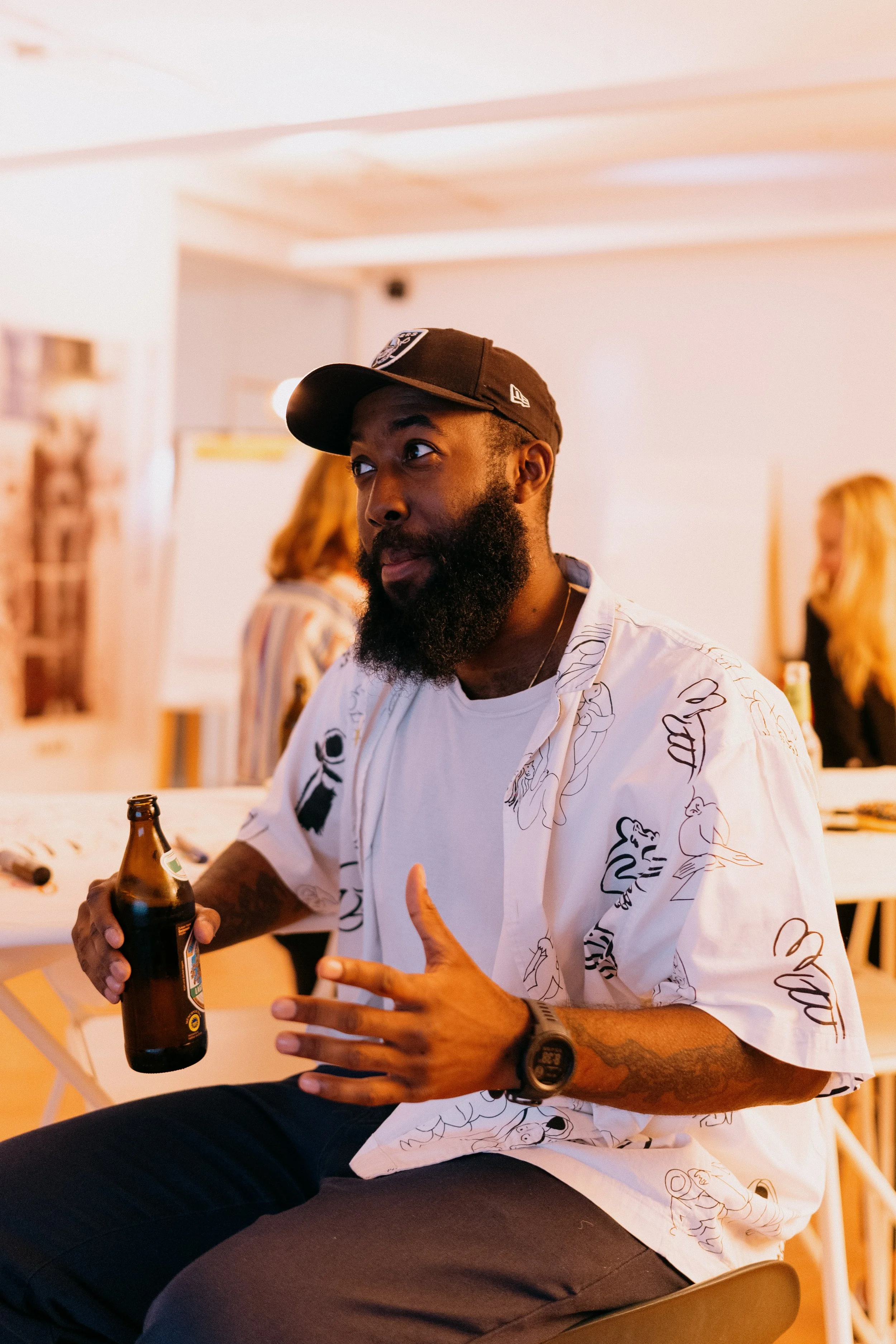 A man with a beard and a baseball cap holding a beer bottle, gesturing with his hand while talking at a social gathering.