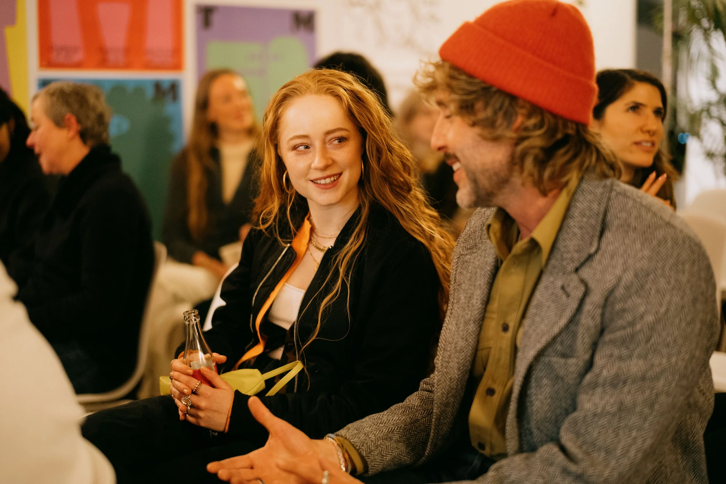 A young woman with long red hair, wearing hoop earrings and a black jacket, sitting next to an older man with long hair, a beard, and wearing a red beanie, gray blazer, and beige shirt, engaged in conversation at an indoor event.