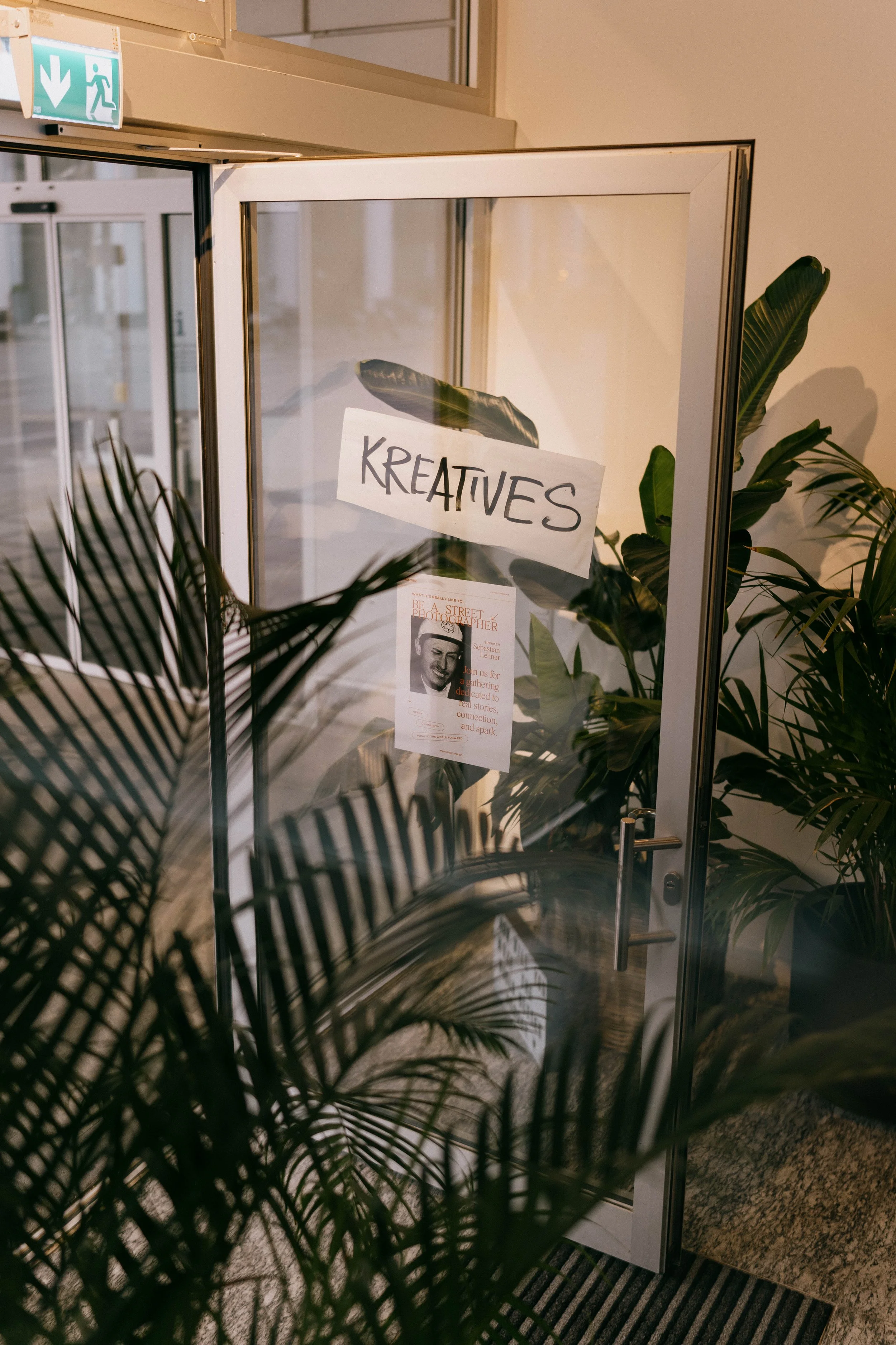 Glass door with a sign that reads 'KREATIVES' taped on it, surrounded by green plants, with a poster visible through the glass door.