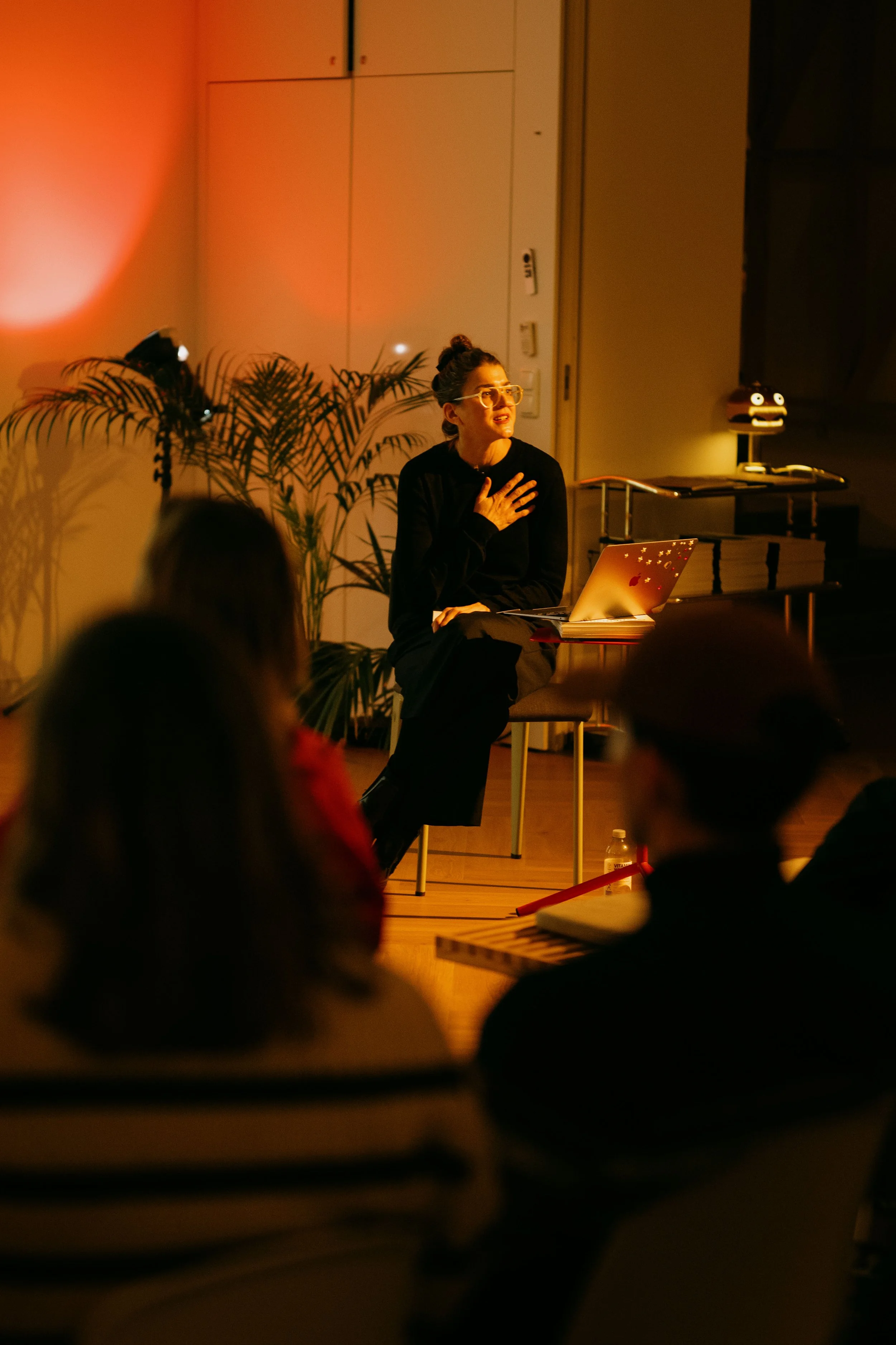 A woman sitting on a chair, speaking or performing in front of an audience, with her hand on her chest, illuminated by warm stage lighting, with a laptop on her lap, and decorative plants in the background.