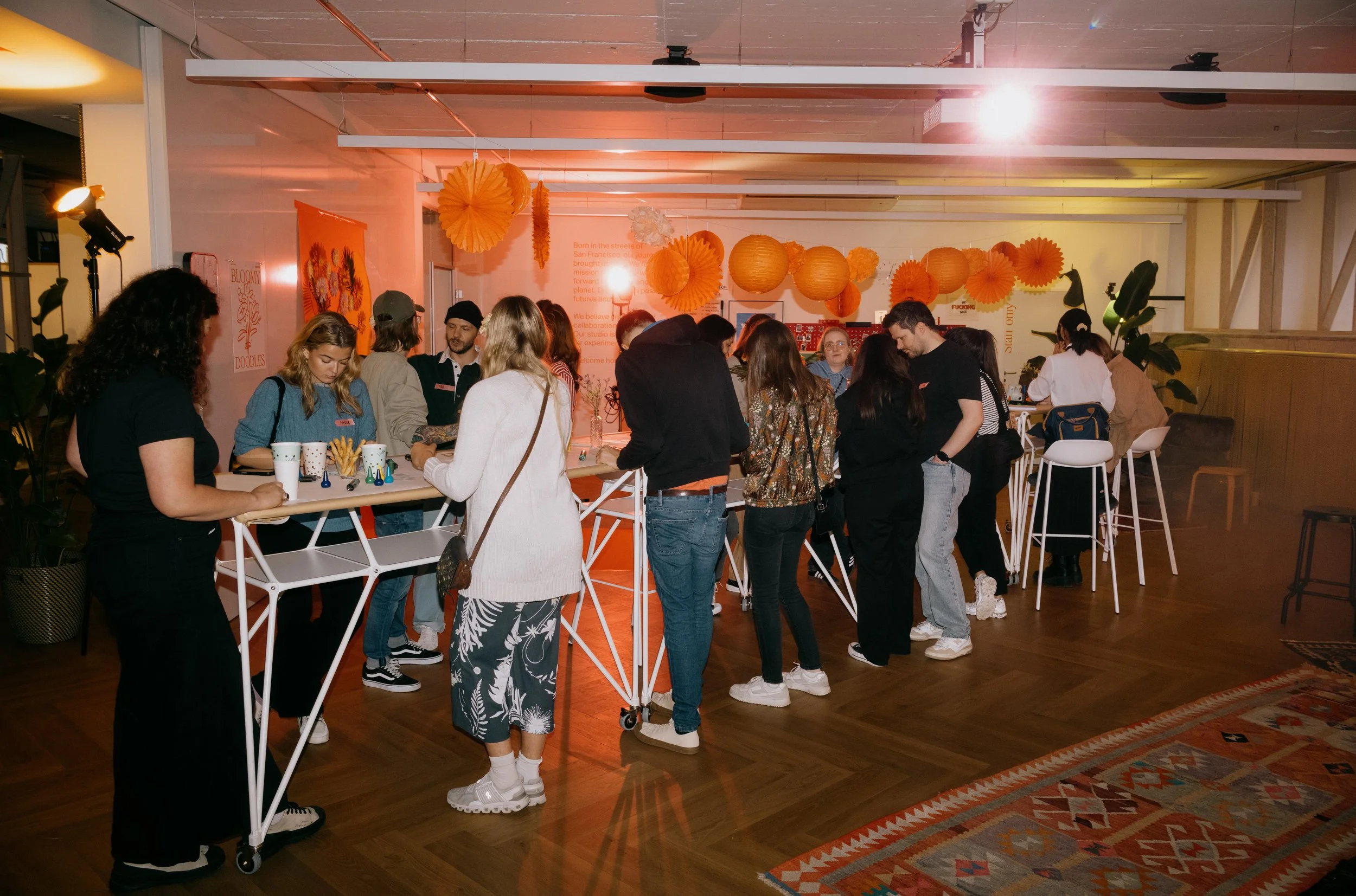 People gathered around a high table in a decorated indoor space with orange paper lanterns and wall decorations, possibly at a social or networking event.