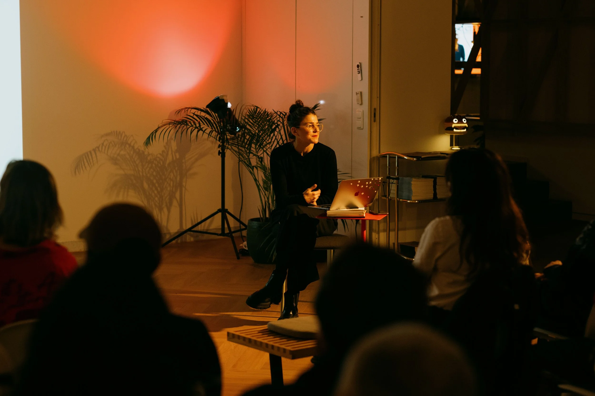 A woman sits in front of an audience, presenting or speaking, in a dimly lit room with warm lighting, a laptop on her lap, and a second screen or projector in front of her.