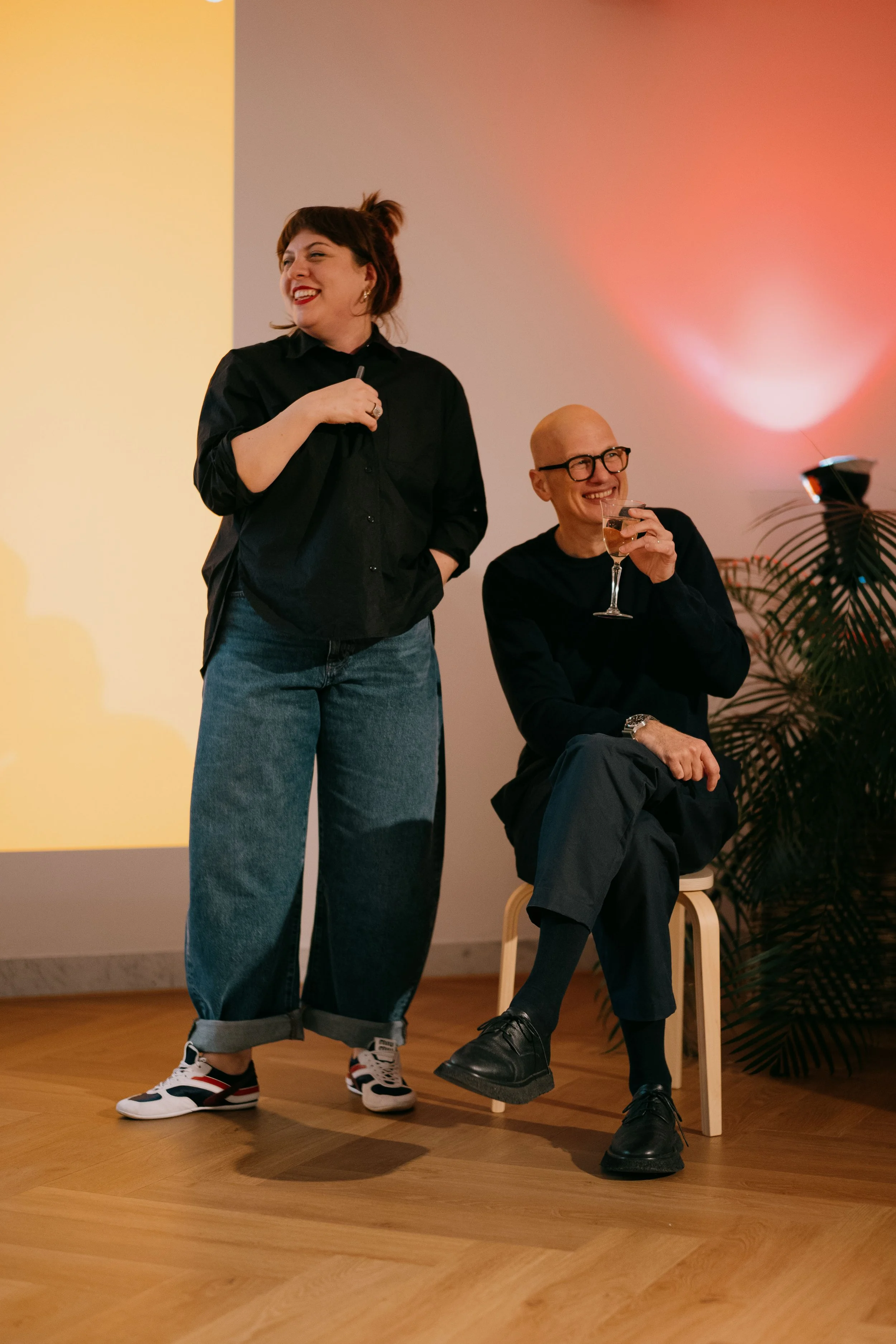 A woman standing smiling in casual attire, and a man sitting on a chair holding a glass of champagne, smiling, in a decorated indoor space.