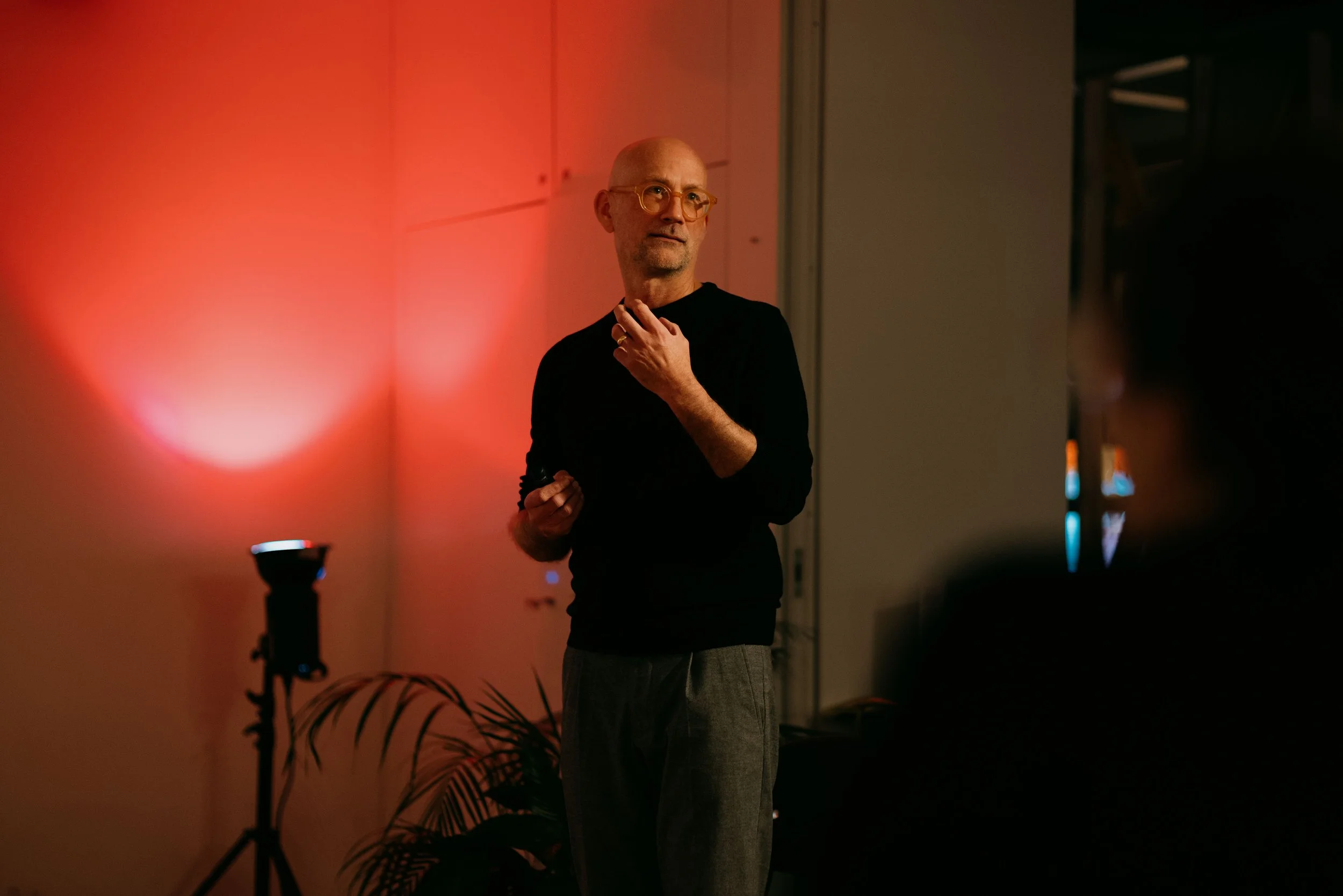 Philipp Osswald, with glasses and a shaved head speaking at an indoor event, illuminated by red lighting on the wall behind him.