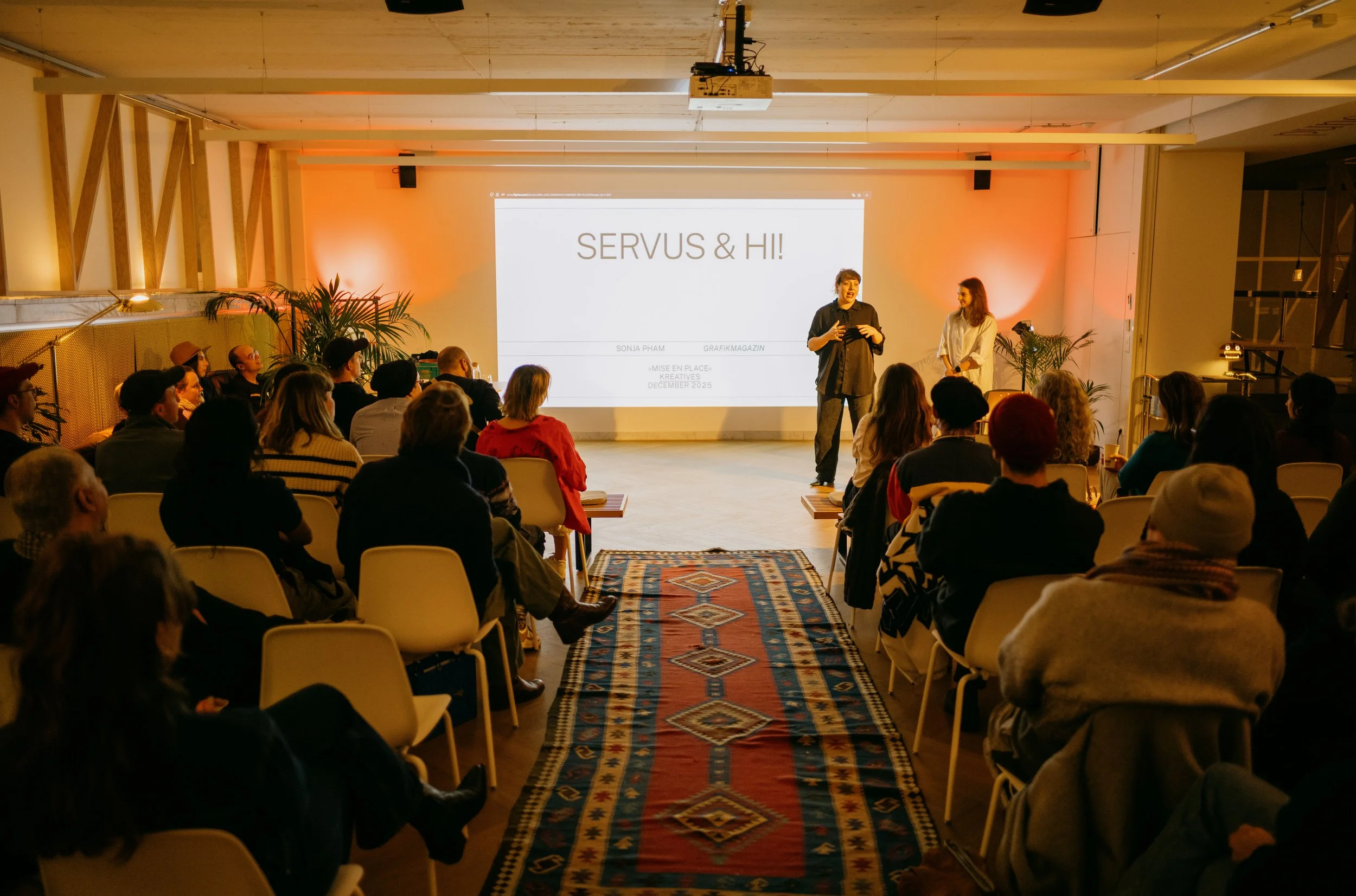 A presentation in a warmly lit conference room with a group of people seated and listening. The speaker is two individuals standing in front of a large screen displaying "SERVUS & HI!". The room is decorated with plants and a patterned rug on the flo