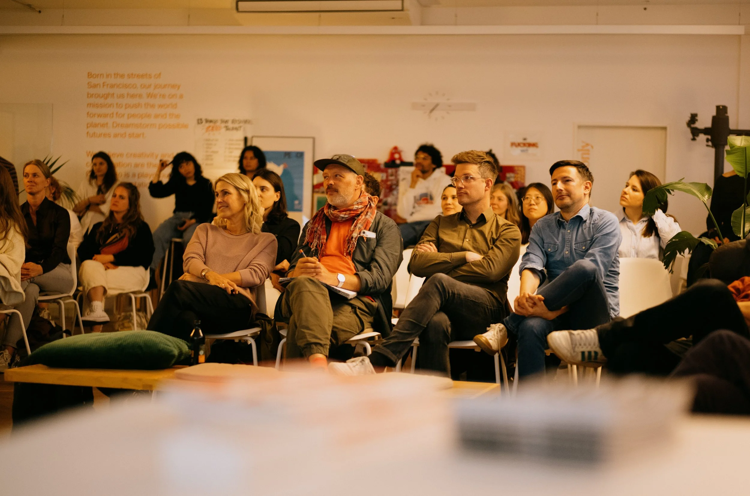 A group of people seated and attentively watching a presentation or event in a well-lit room with a white wall in the background.