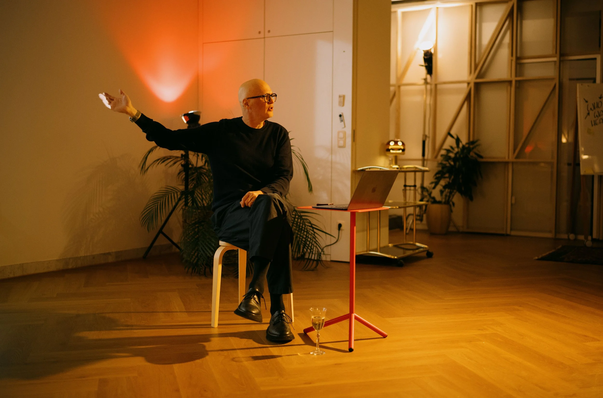 Person with short white hair, glasses, wearing black clothing, sitting on a white chair, gesturing with-arm extended, in a warmly lit room with plants and wooden flooring, a laptop on a small red table, and a glass of wine on the floor.