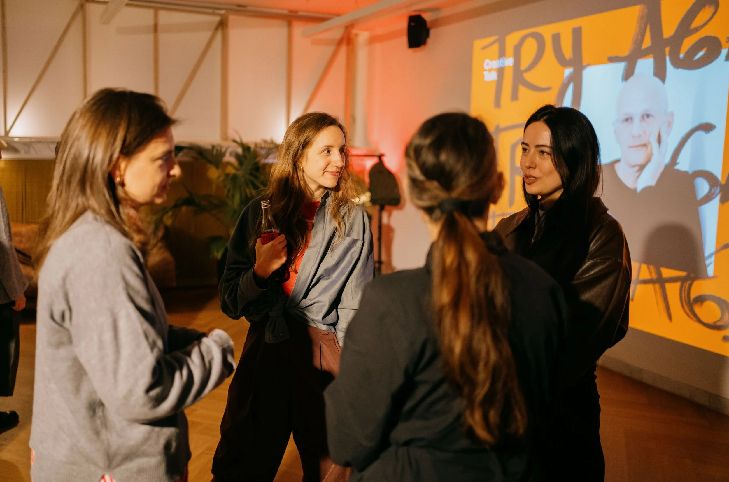 A group of five women talking and socializing at an indoor event with colorful artwork and lighting in the background.