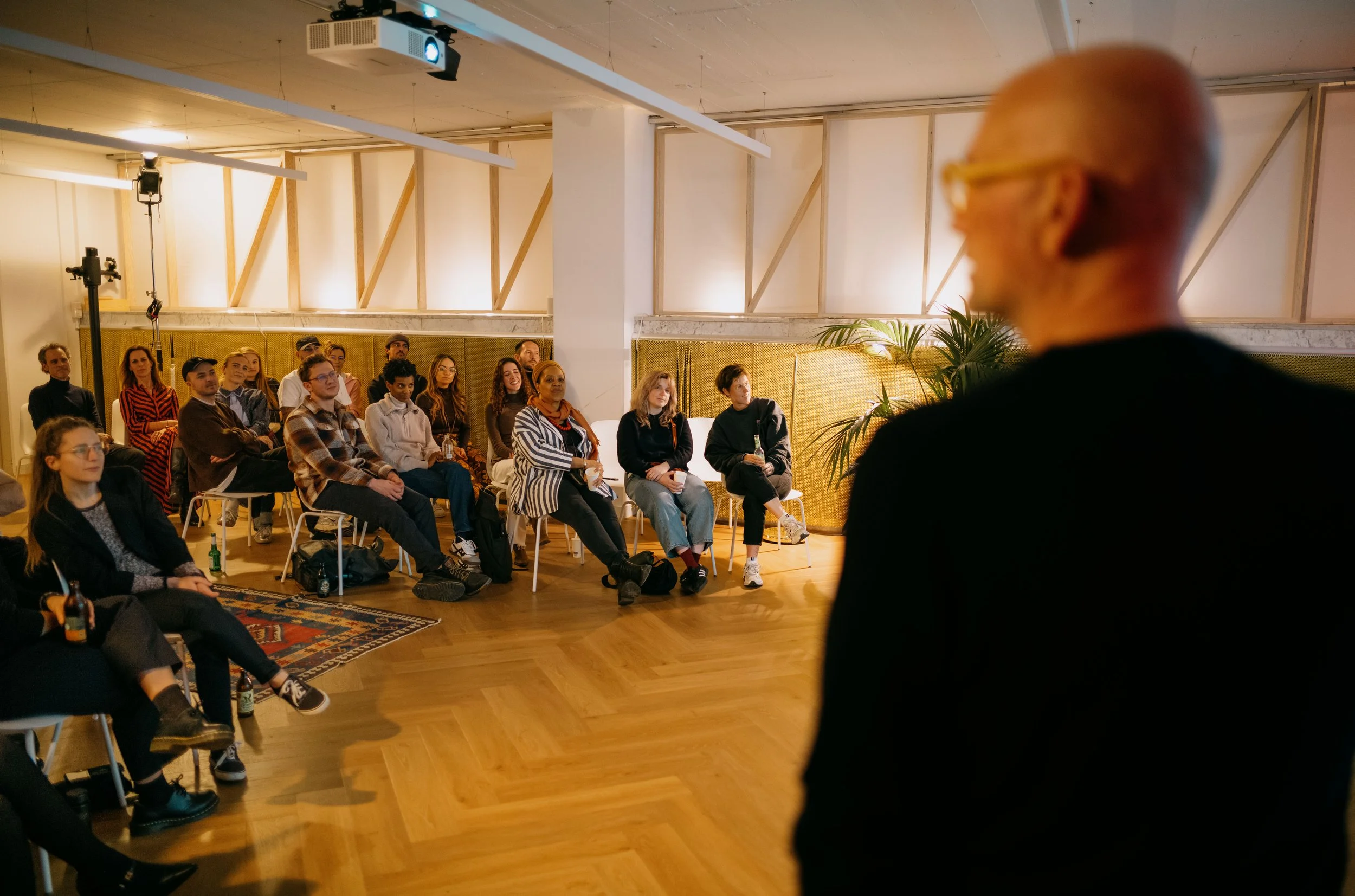 An older man with glasses and a black jacket speaking to an attentive audience seated in a room with wooden floors, plants, and decorative walls.