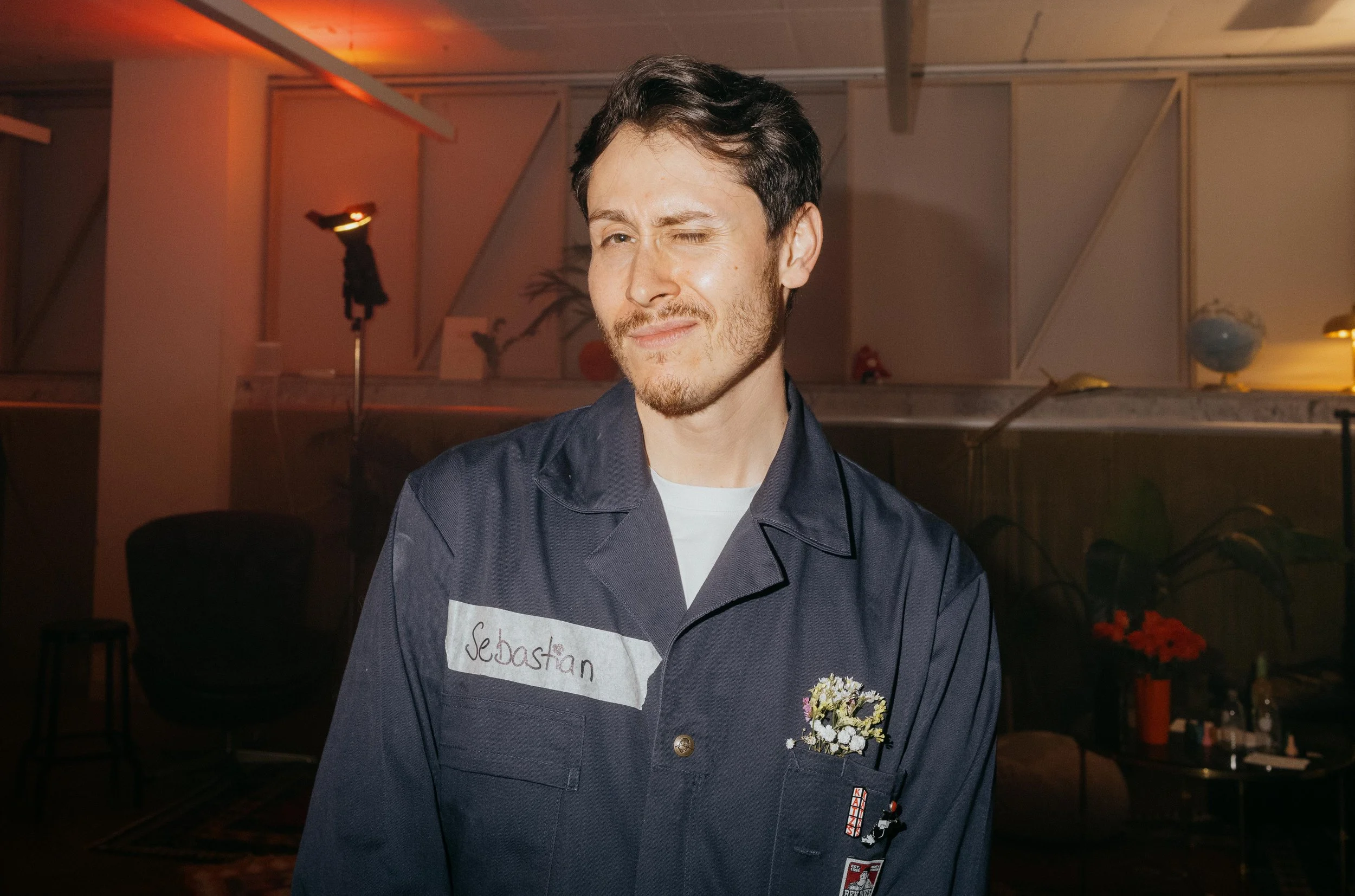 A man with dark hair and facial hair, wearing a navy blue work shirt with a name tag reading "Sebastian" and a small patch on the pocket with flowers. He is winking and has a slight smile, standing in a room with warm lighting, chairs, and decorative