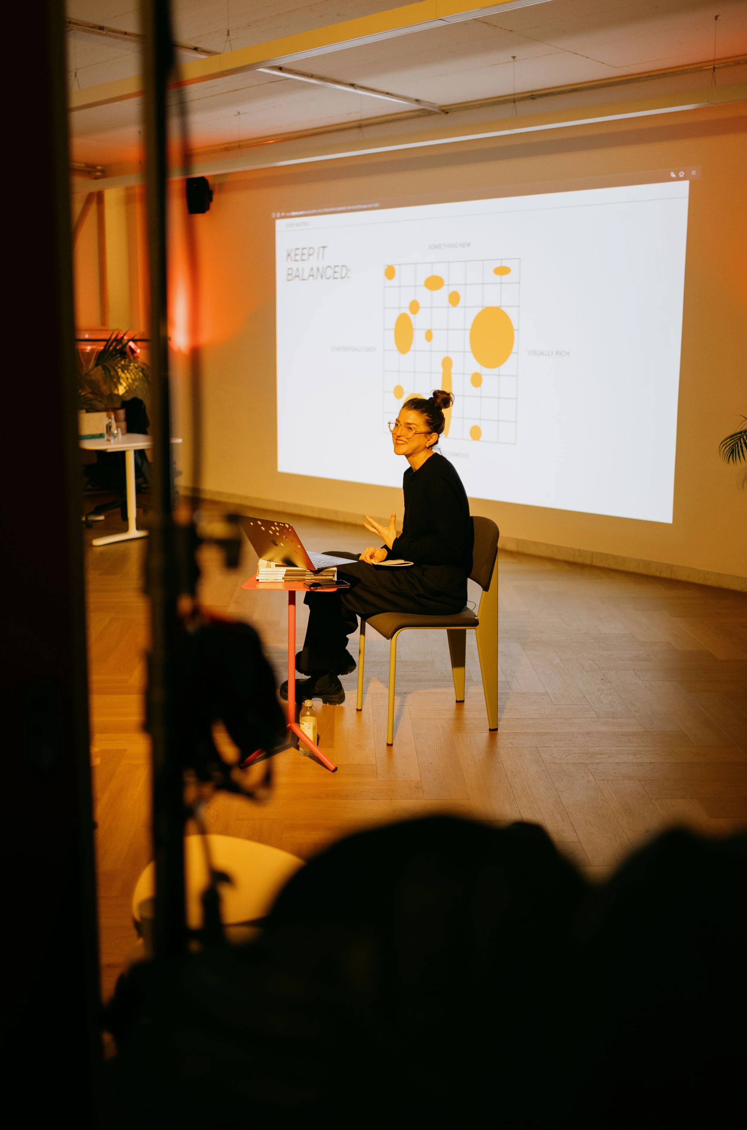 A woman sits on a chair on a stage, smiling and animated while giving a presentation. A large screen behind her displays a graph with circles of varying sizes and colors, labeled 'Keep it Balanced' with axes indicating 'Visual Rich' and 'Something Ne