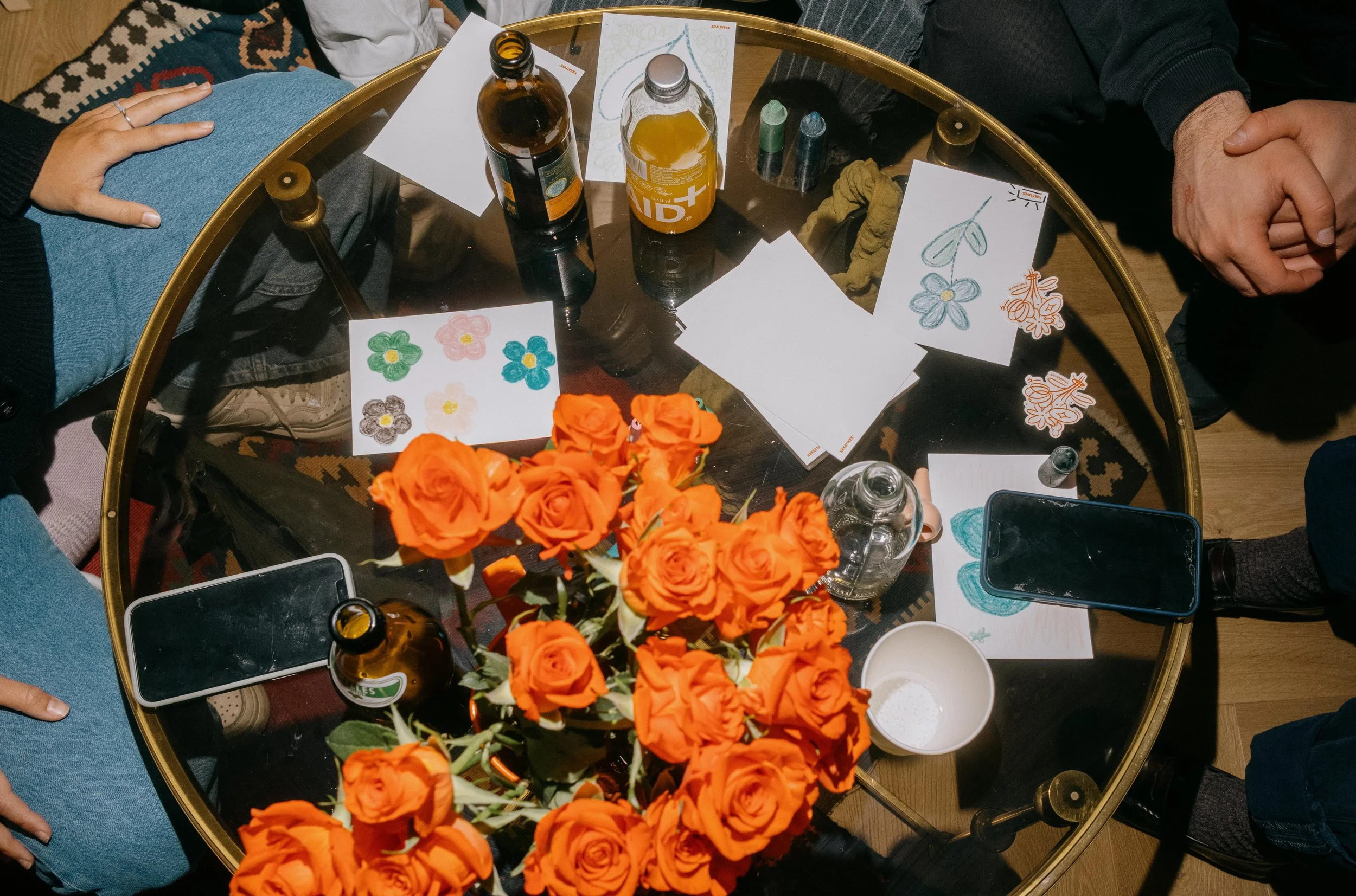A round glass table with orange roses in the center, surrounded by three people, one with a hand resting on their lap and another with folded hands. The table holds bottles of beverages, drawing papers with flower sketches, markers, a water bottle, a