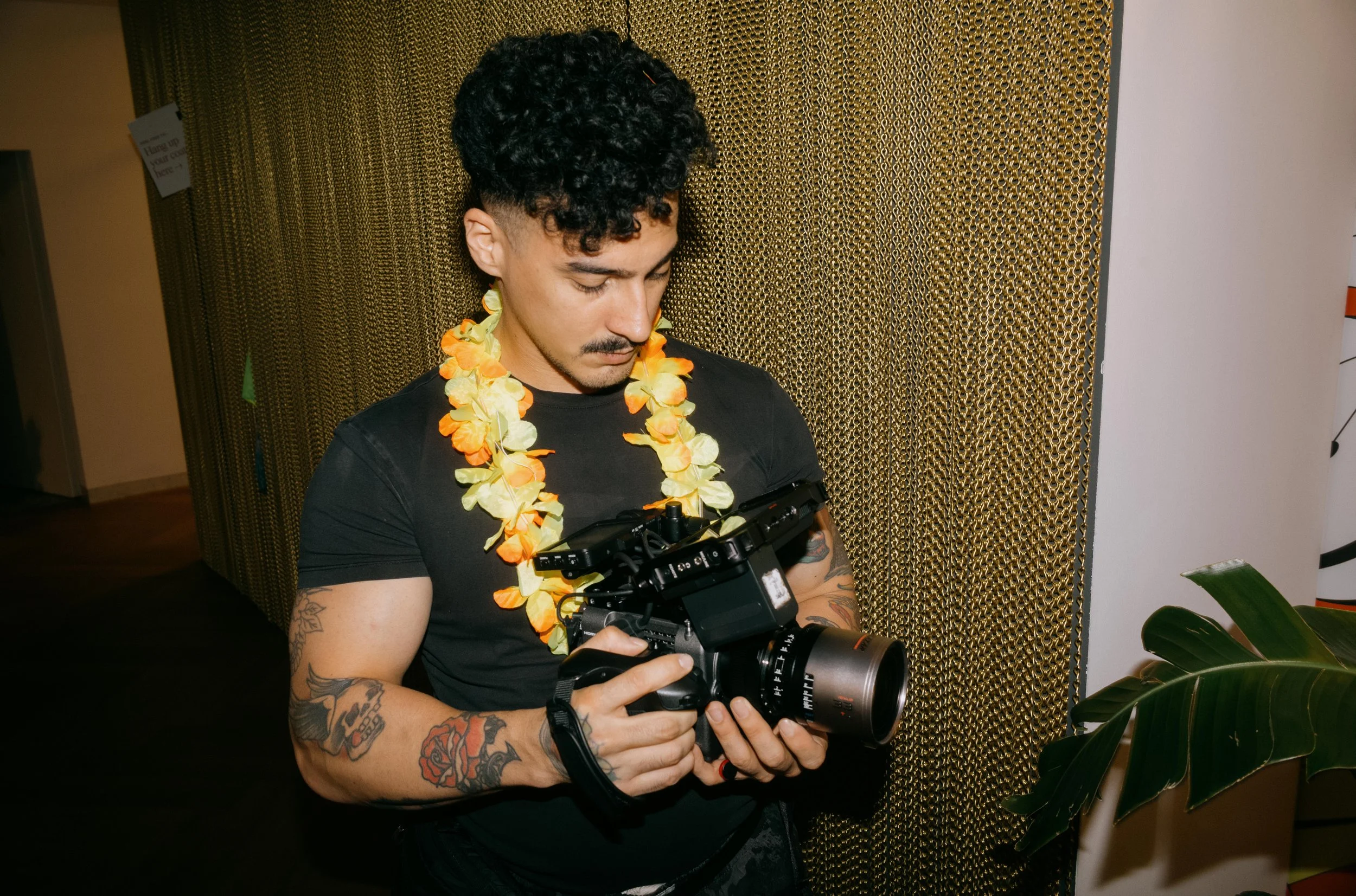Man with tattoos wearing a black shirt and leis, holding a camera, standing indoors near a patterned gold wall.