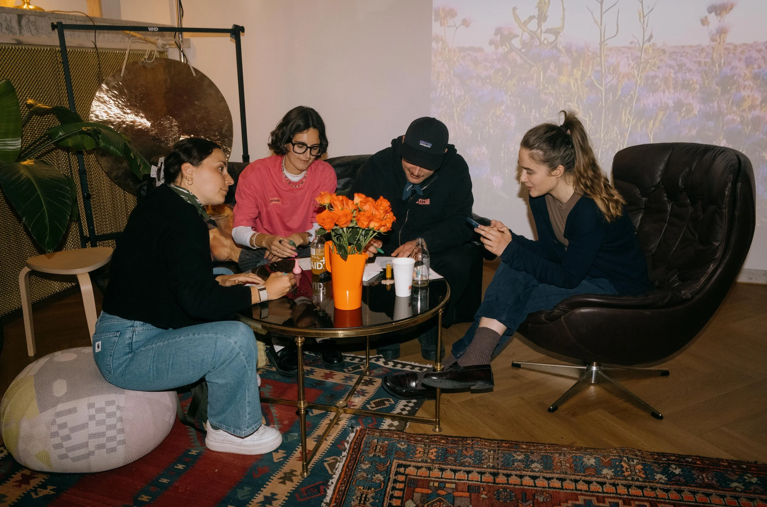 Five young adults gathered around a coffee table with a vase of orange flowers, engaging with their phones and drinks in a cozy indoor setting.