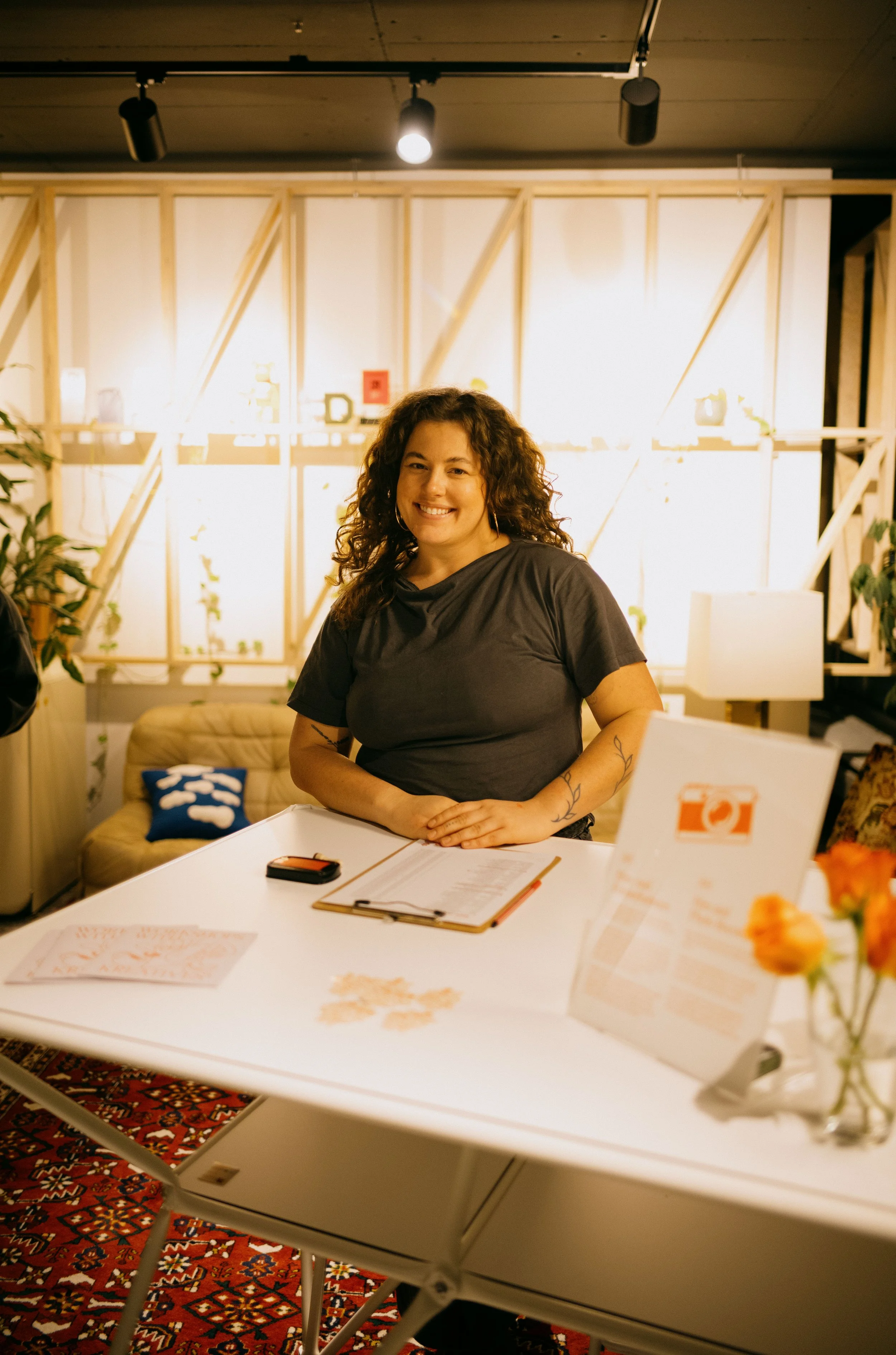 A woman with curly brown hair and a tattoo on her arm, wearing a black t-shirt, smiles while standing behind a table with a clipboard, a smartphone, and some papers, in a warmly lit room.