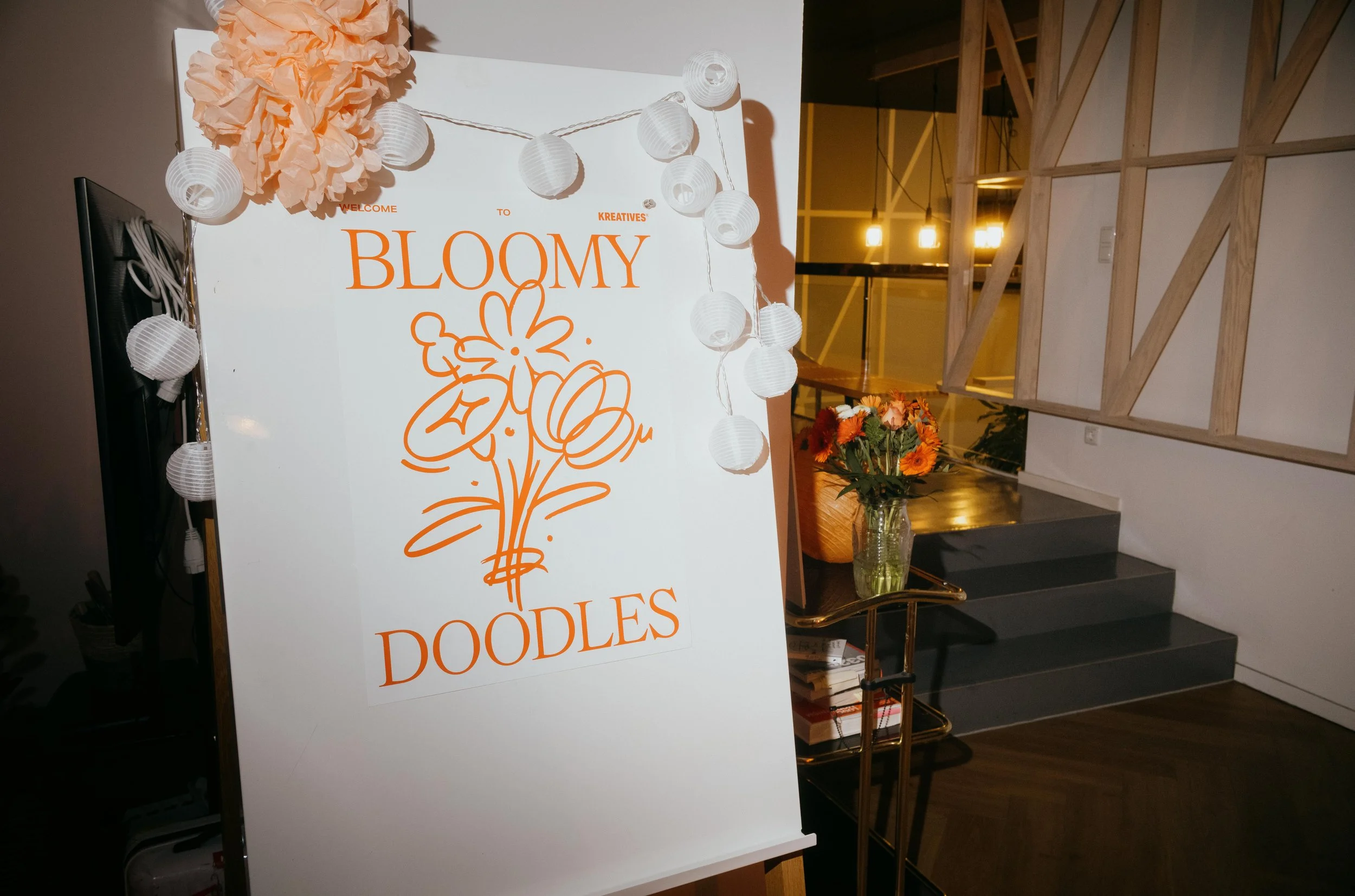 Event sign with orange text reading 'Welcome to Bloomy Doodles' and a floral illustration, decorated with white paper lanterns. A vase of orange flowers is on a small cart nearby, with wooden steps and warm lighting in the background.