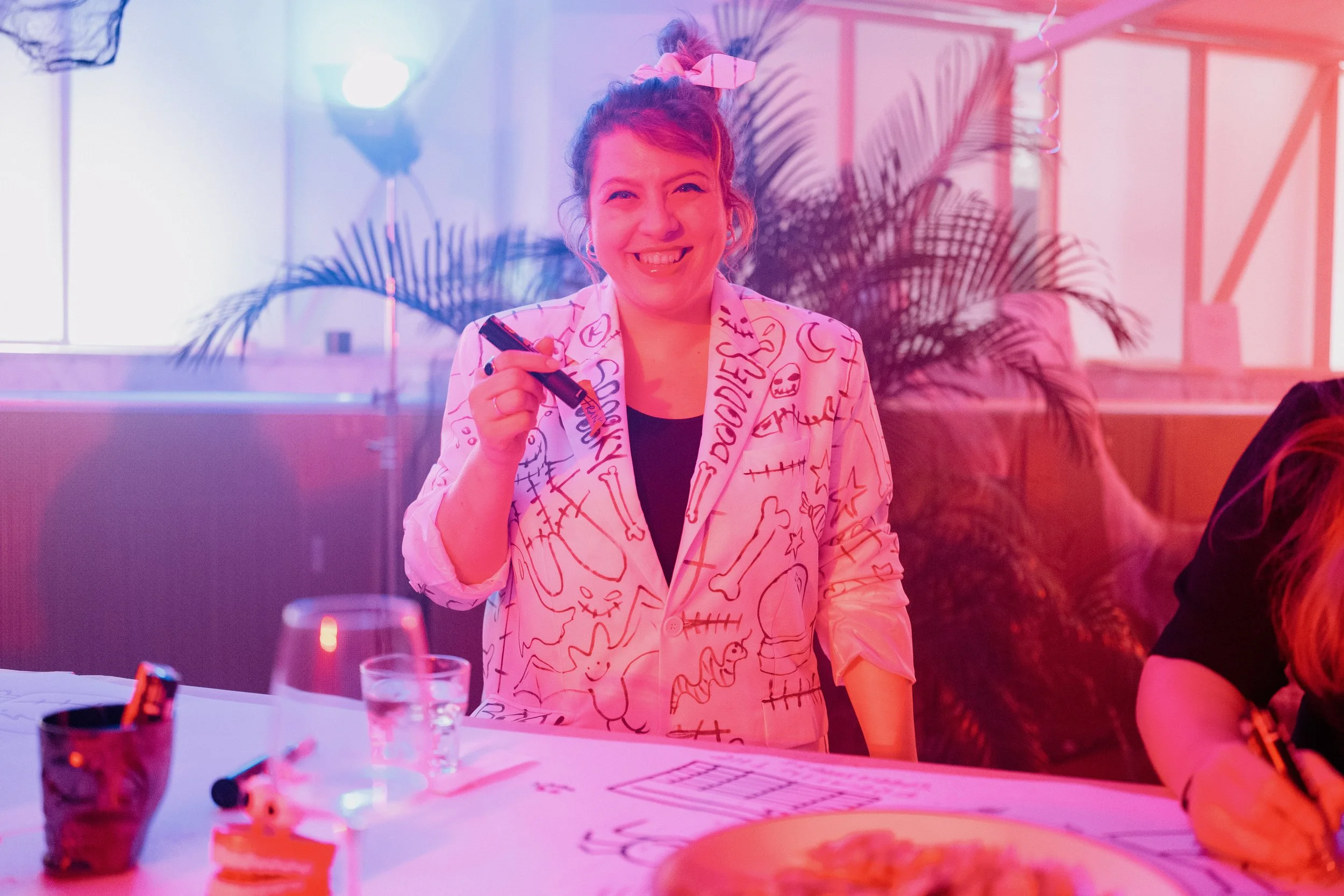 A woman smiling and holding a black marker, wearing a white blazer with colorful doodles and words, standing at a table with drinks and paper, with pink and purple lighting and plant in the background.