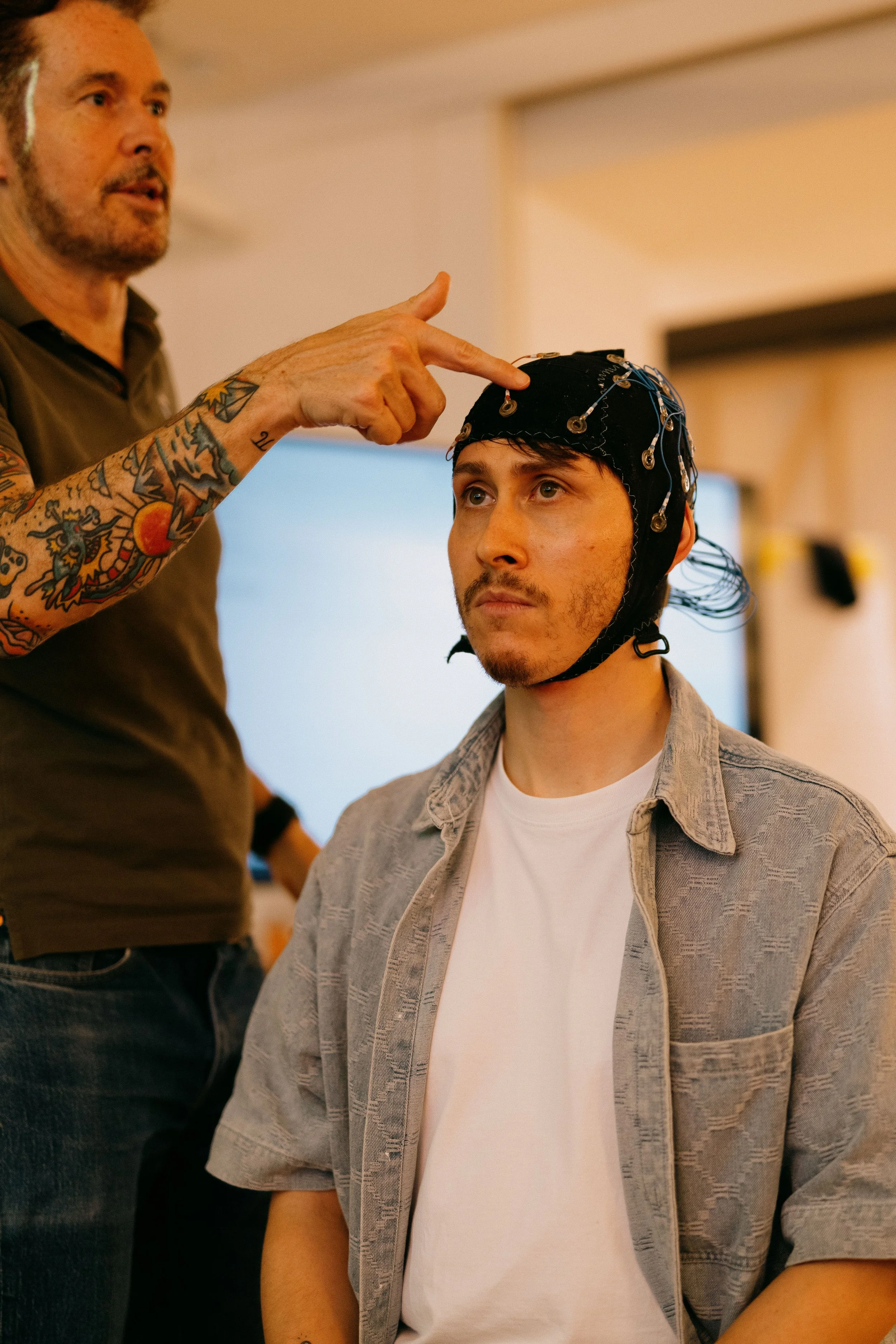 A man wearing a cap with electrodes connected to a neuroscience device, sitting in a room while another man adjusts or explains the setup.