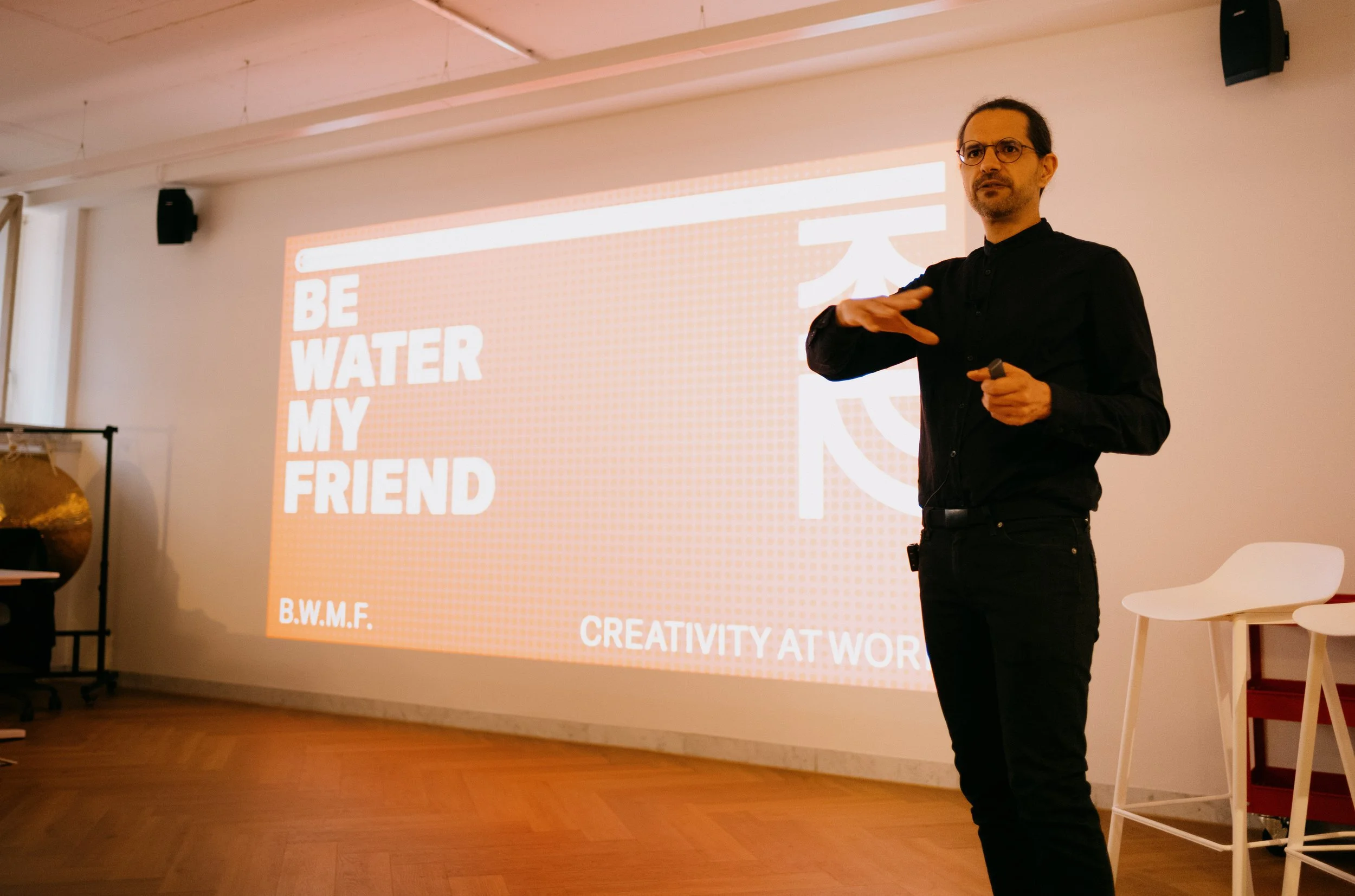 Man giving a presentation in a room with a large screen displaying the text 'BE WATER MY FRIEND' and 'CREATIVITY AT WORK' in the background
