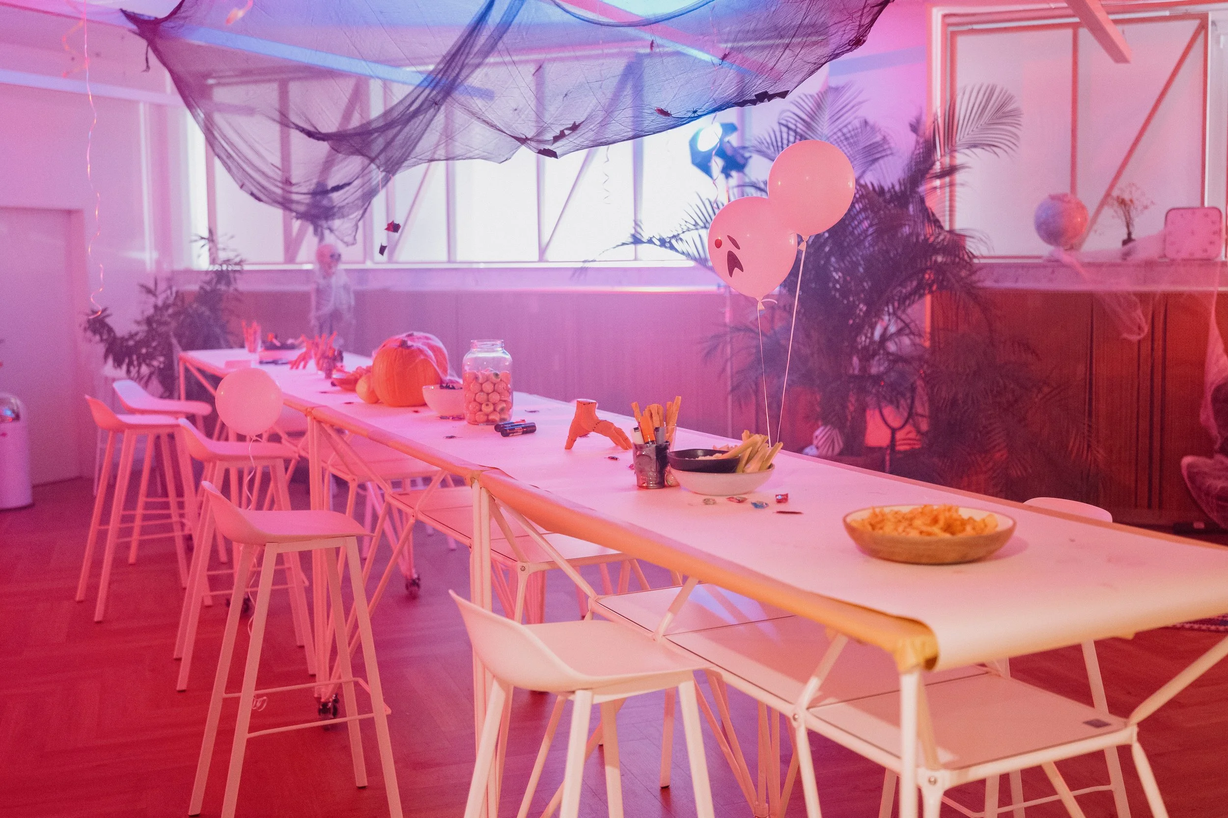 Party setup with a long white table decorated with balloons, including two with sad faces, assorted snacks, and Halloween decorations in a room with pink lighting, plants, and spider webs.
