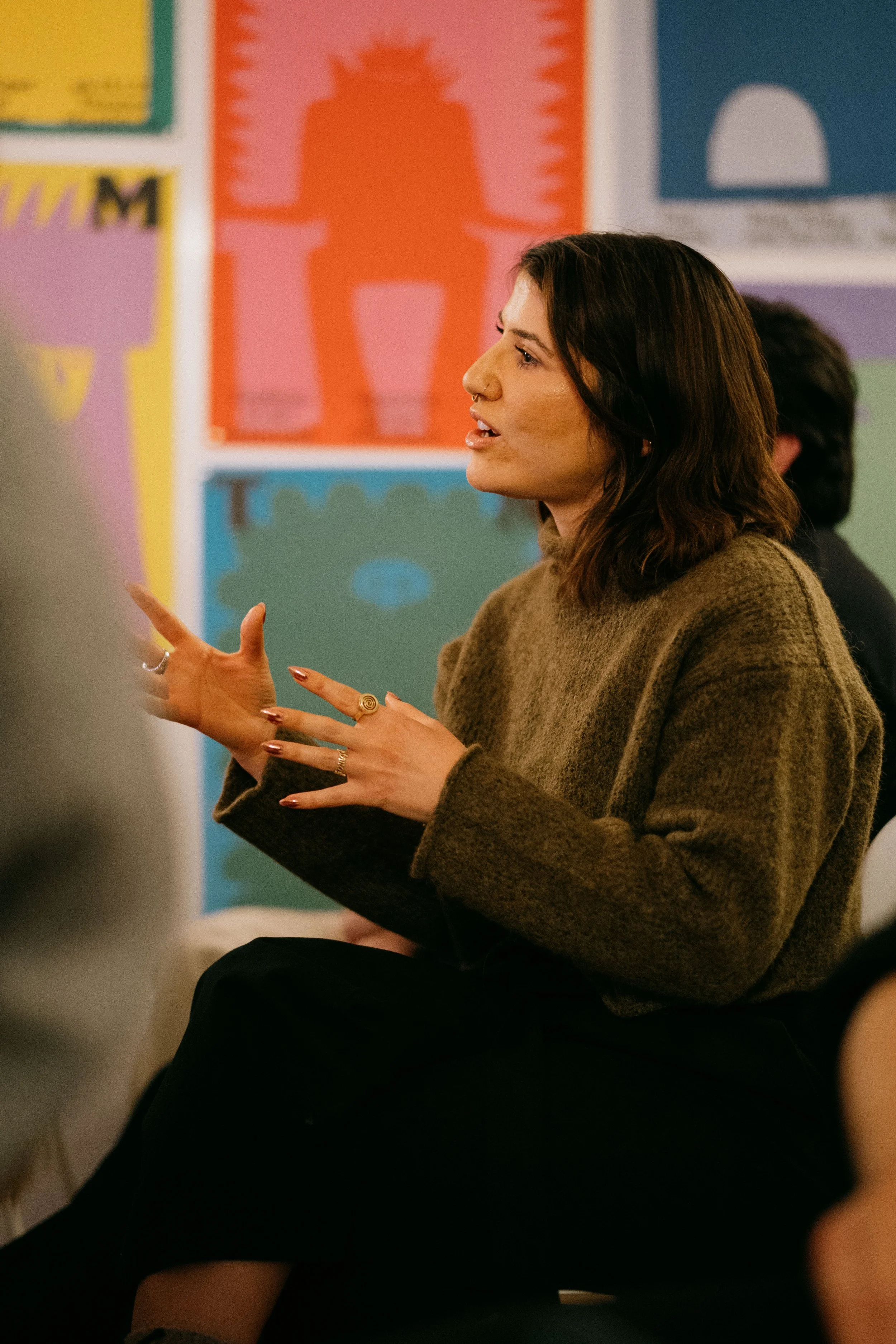 A woman with shoulder-length dark hair and a nose ring is speaking in a room with colorful posters or artwork on the walls.