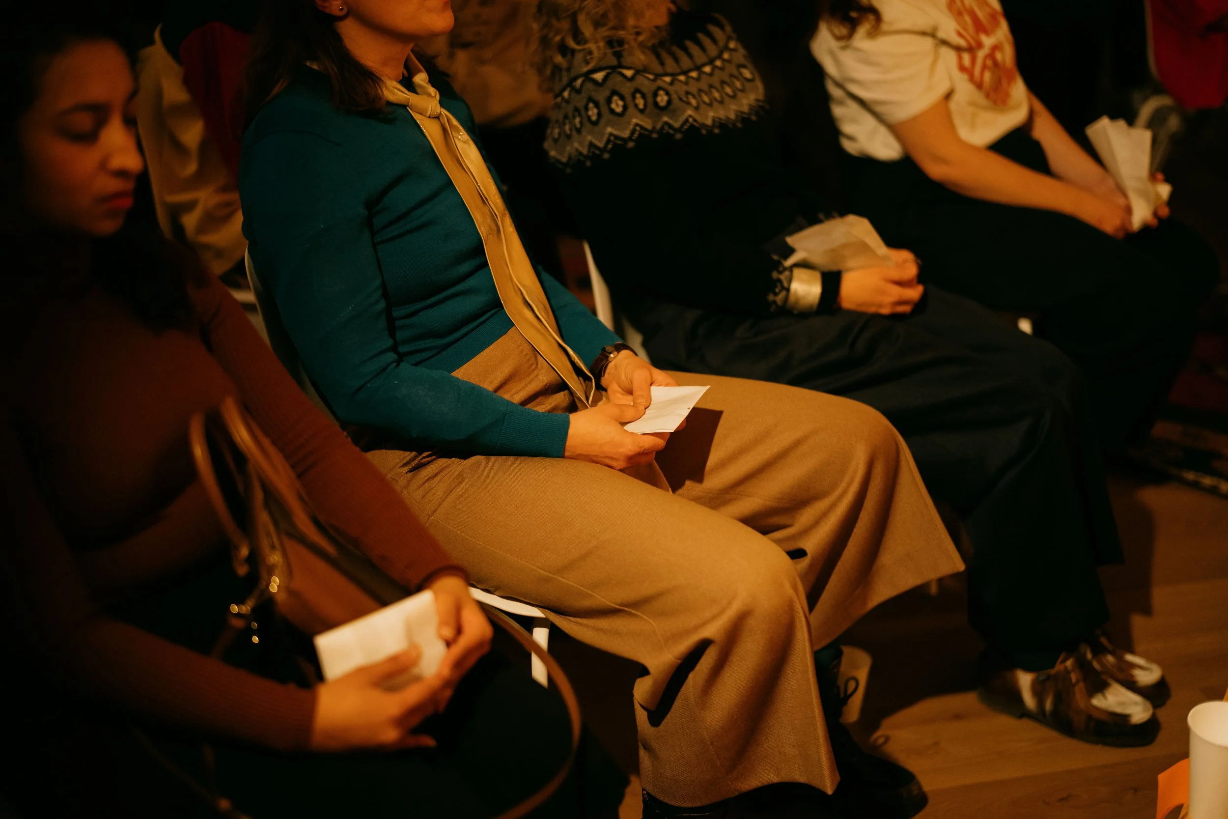 A group of people sitting on chairs at a conference or meeting, holding papers or notes, with a focus on a woman in a teal sweater and beige pants.