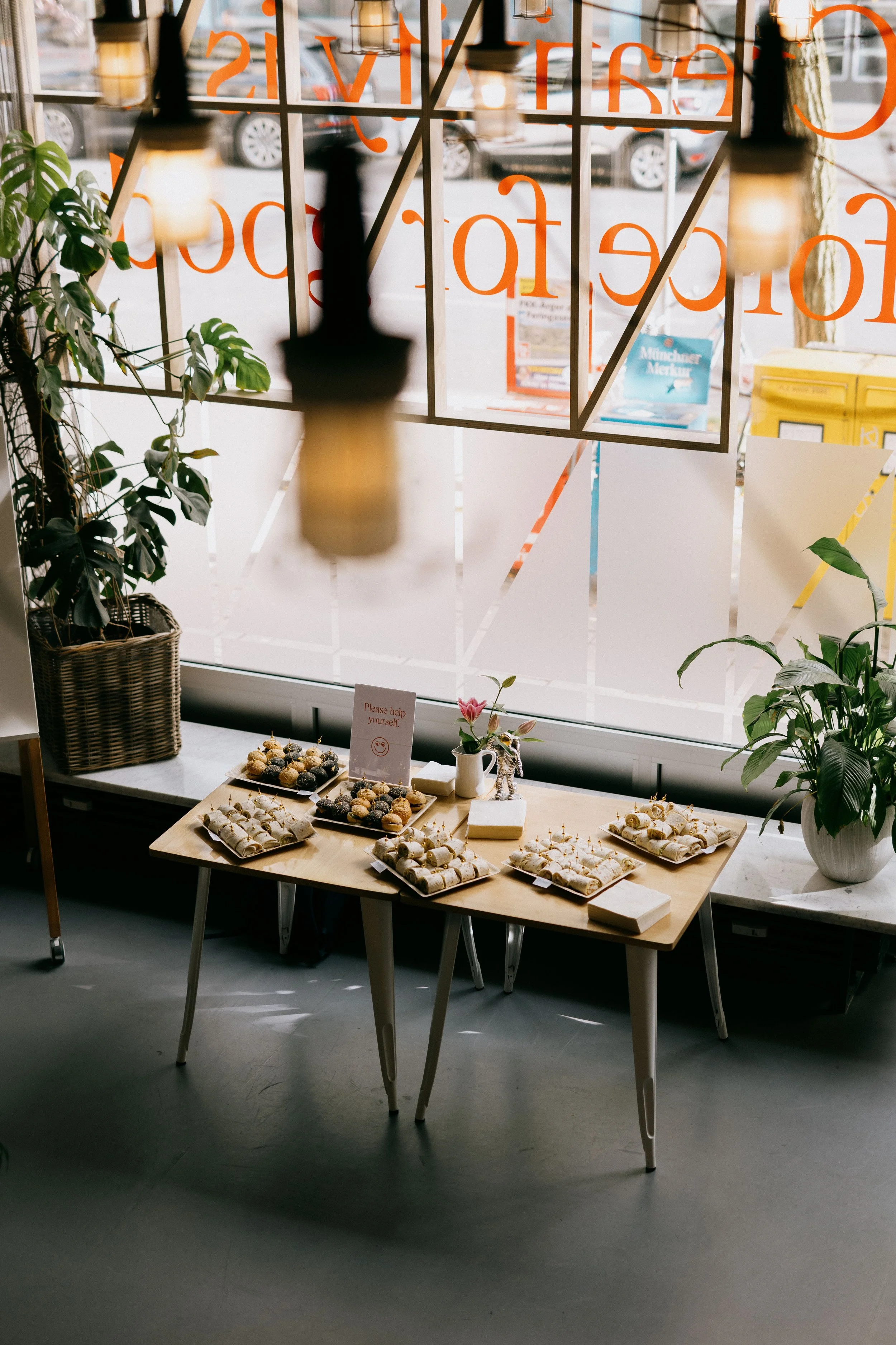 A table with a variety of baked goods and a sign that reads "Please help yourself." The table is near a large window with potted plants on either side.