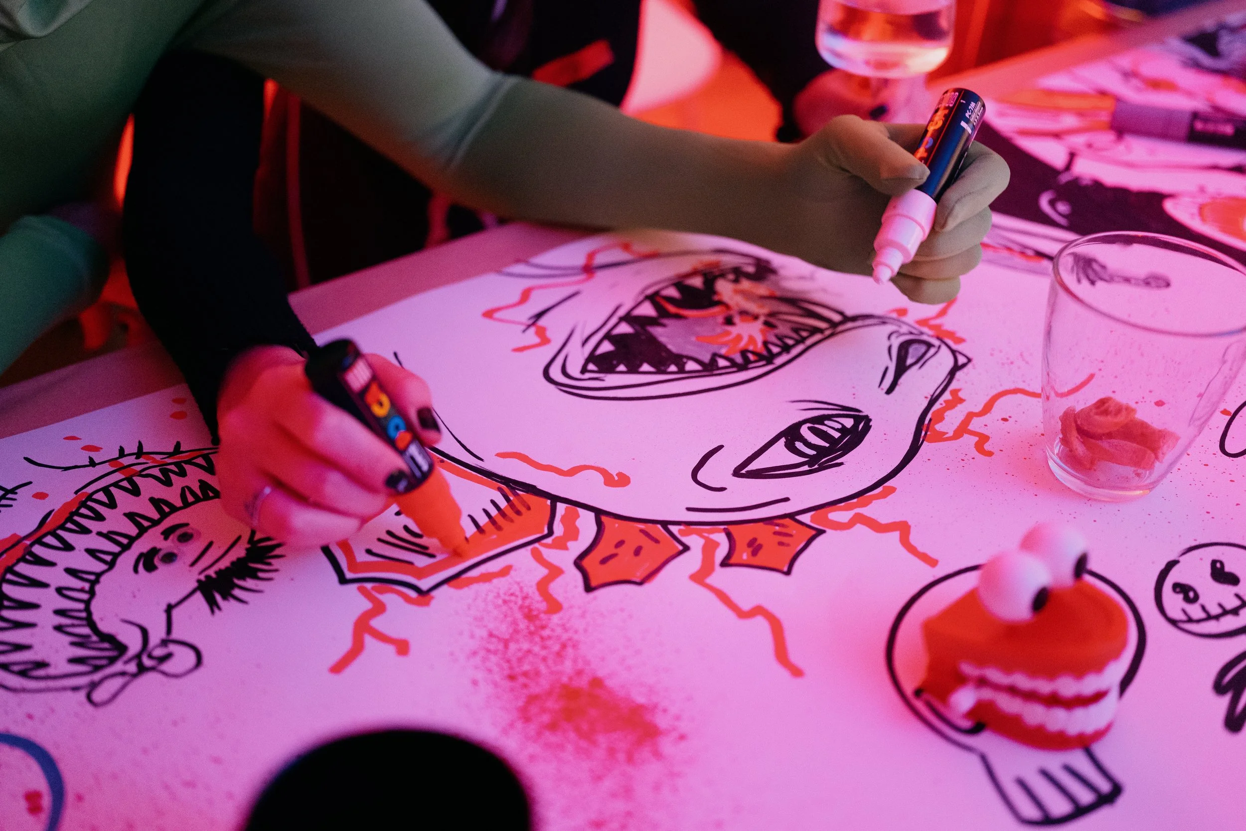 Person drawing a colorful, stylized face on a white poster with markers, surrounded by paint supplies and a glass with a piece of bread.