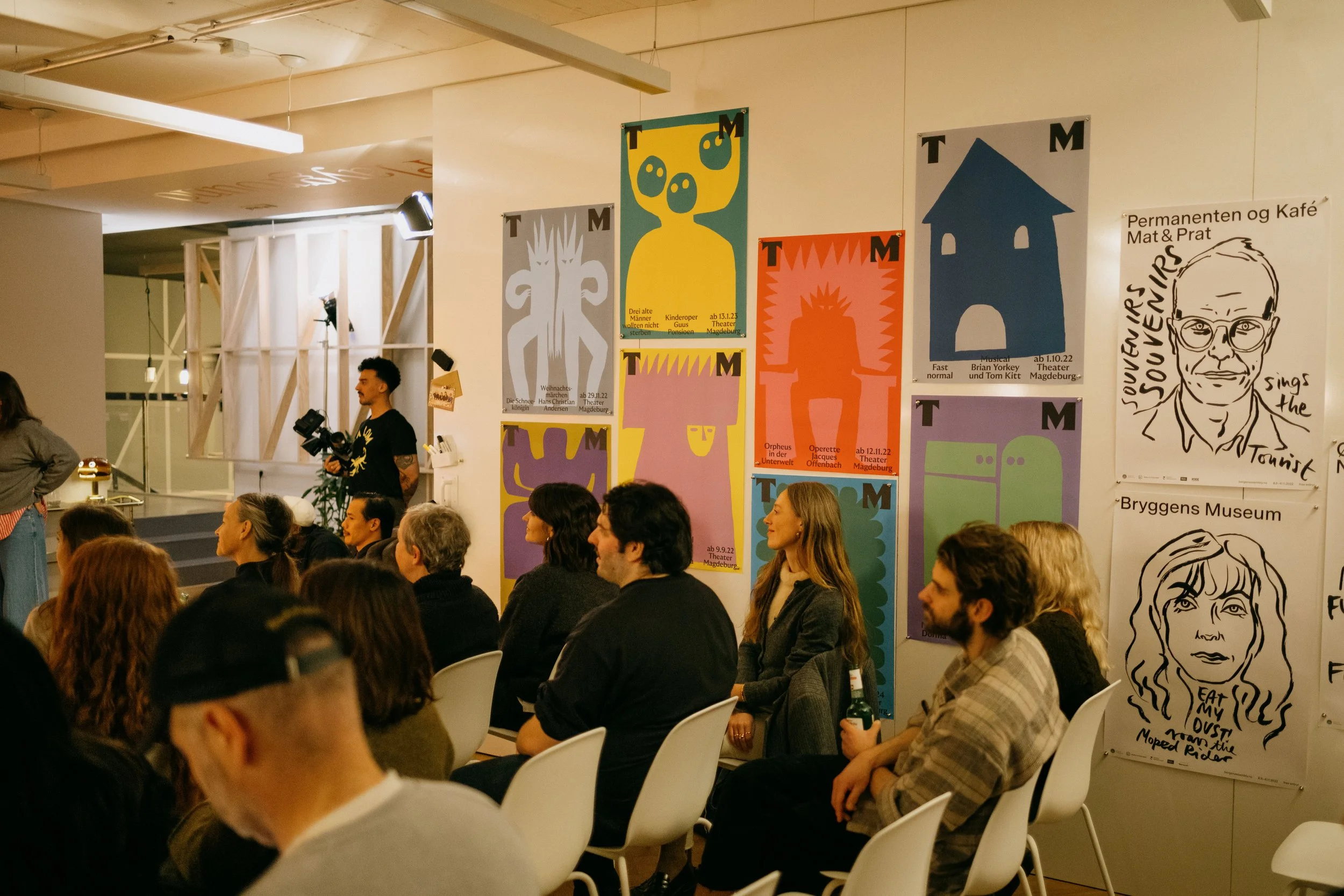 Audience seated in a room during a presentation, with colorful posters on the wall behind them, including abstract art and sketches of people's faces.