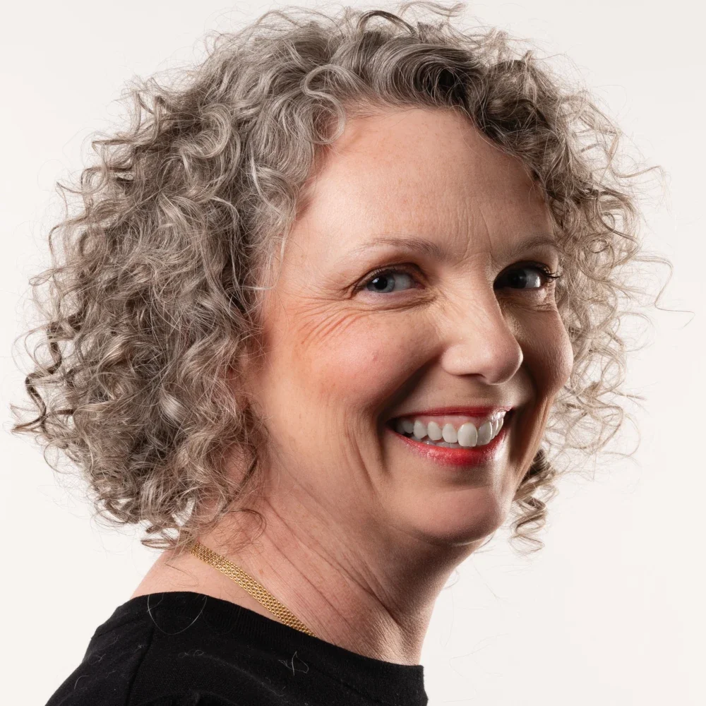 A smiling middle-aged woman with curly gray hair, wearing a black top and gold necklace, against a plain white background.