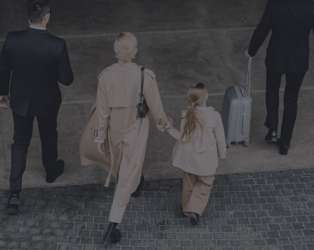 A family of four, including two adults and two children, walking together on a sidewalk at an airport, with the children holding hands, and one adult pulling a rolling suitcase.