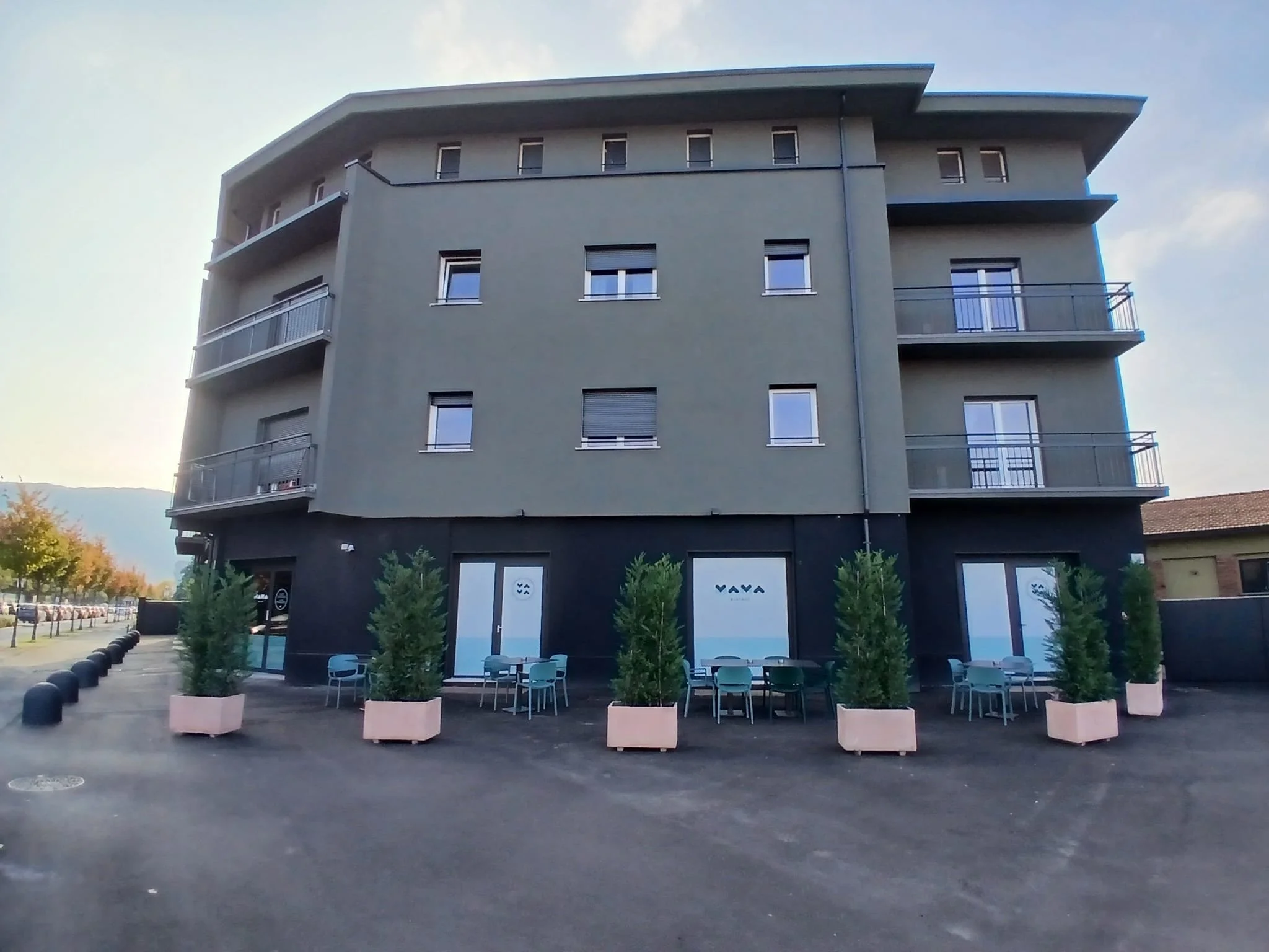 Edificio moderno con negozio e terrazze con sedie e piante davanti, cielo sereno.