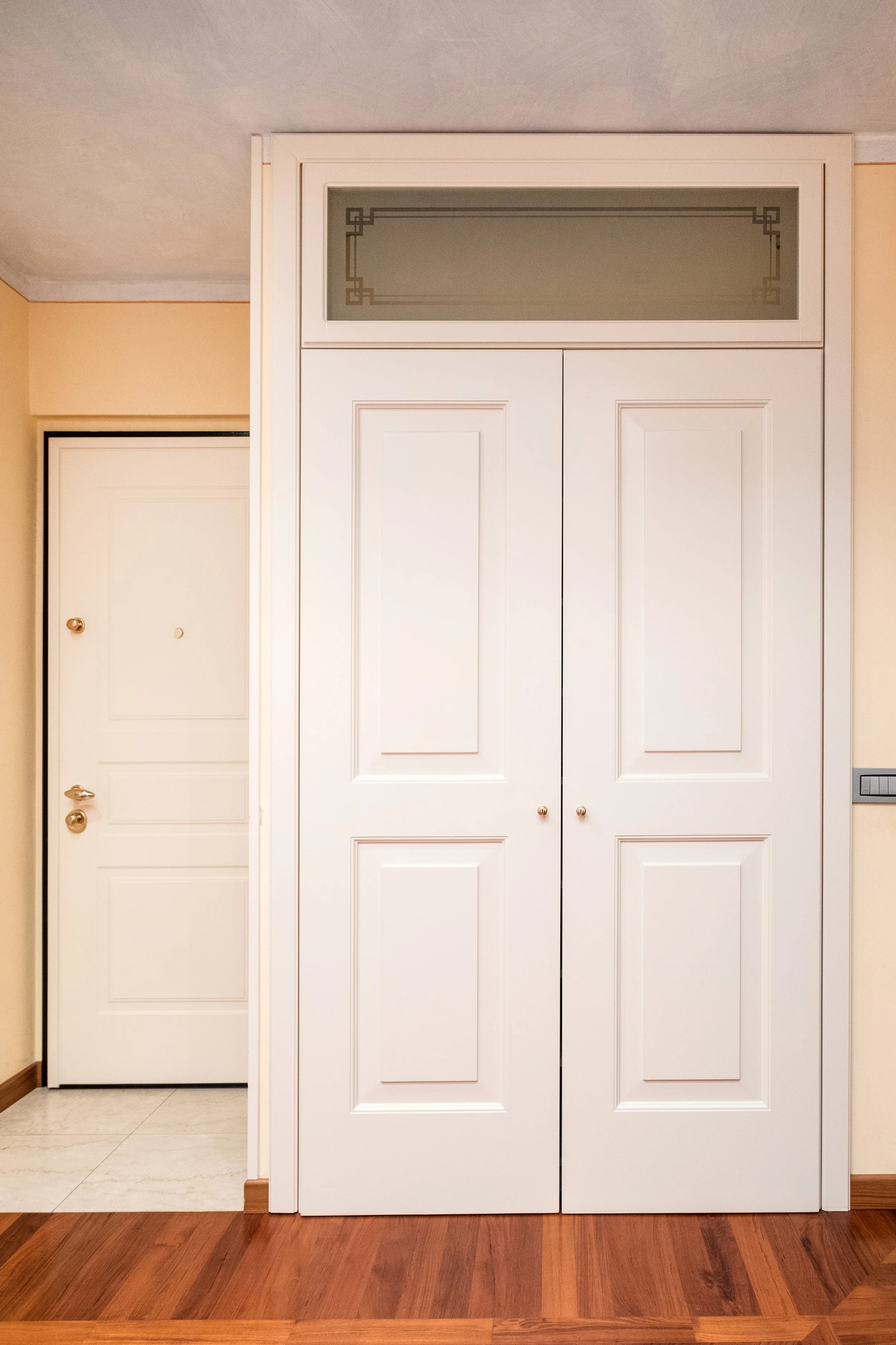 Porta bianca a due ante con maniglie rotonde, accanto a una porta di ingresso con maniglia e citofono, in un ambiente con pavimento in parquet.