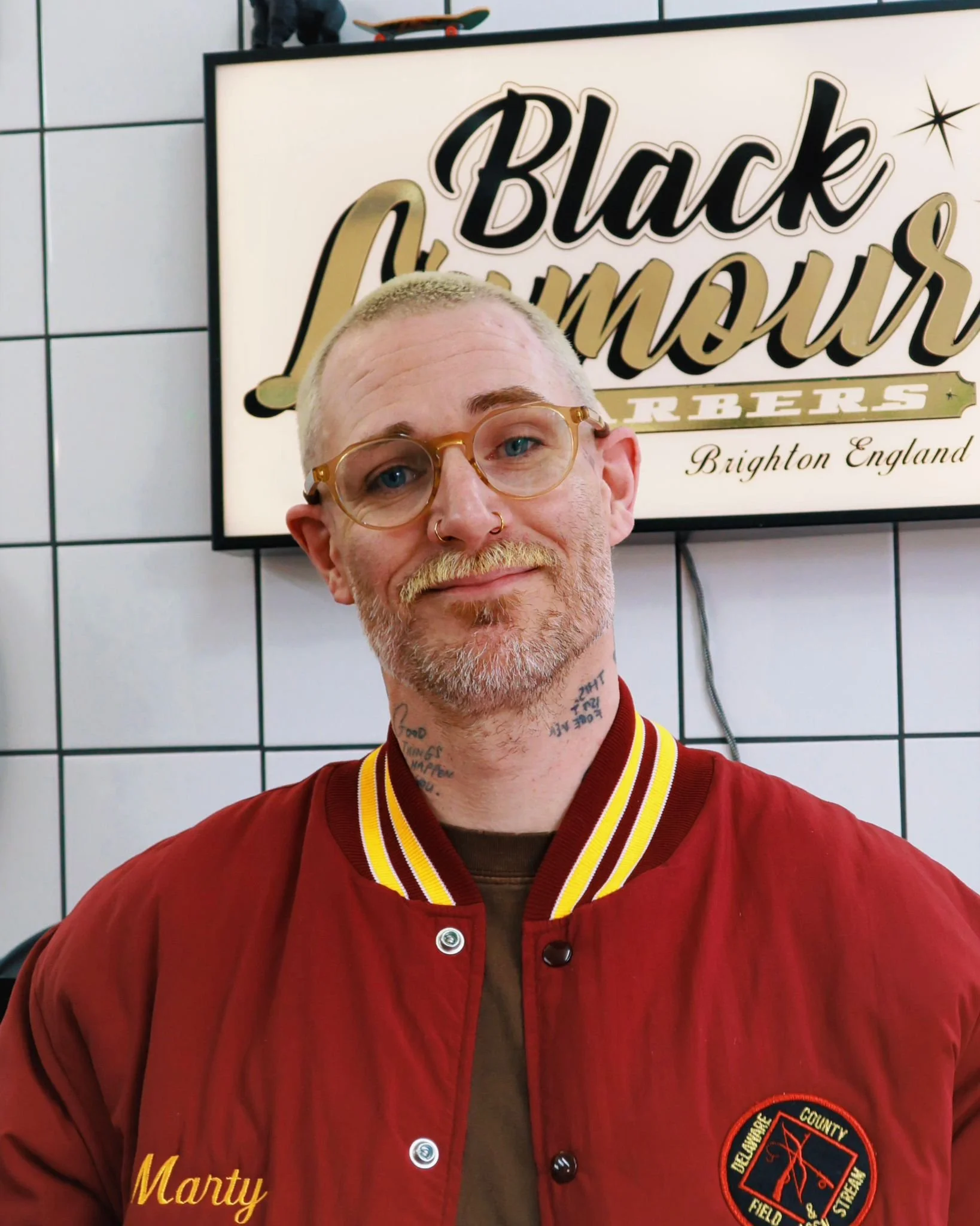 A man with glasses and a beard, wearing a red jacket with 'Marty' embroidered on it, standing in front of a sign that reads 'Black Lumberjurs Brighton England.'