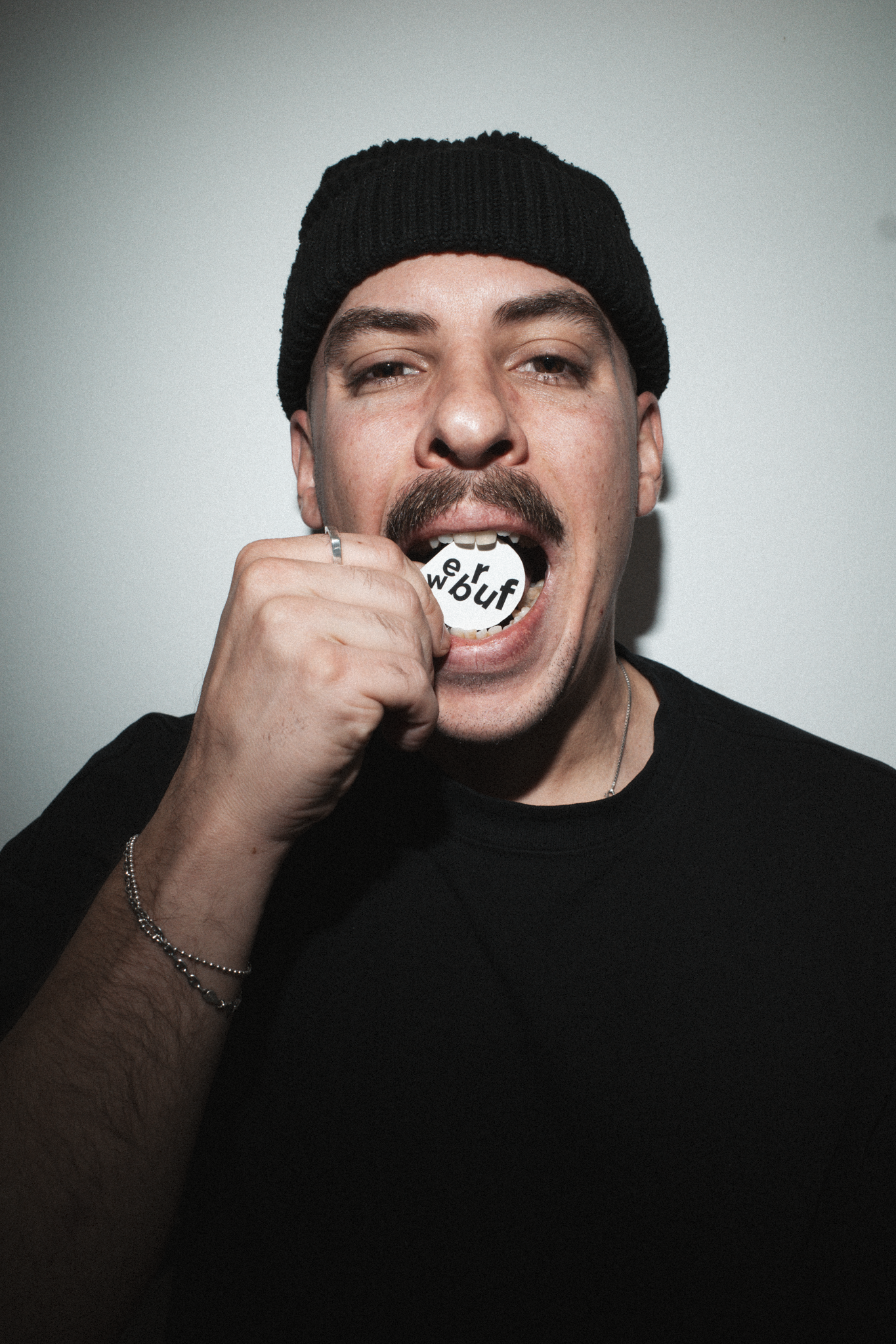 Junger Mann mit schwarzer Mütze, der eine runde Karte mit den Buchstaben 'er buf' im Mund hält, vor einfacher grauer Wand.