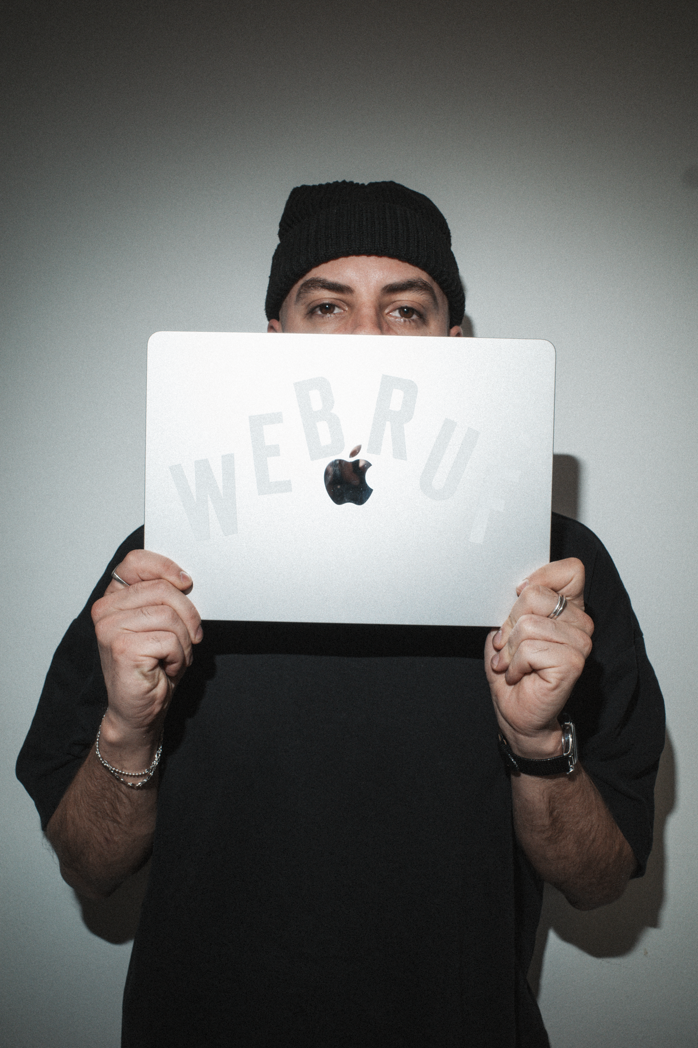 Person trägt einen schwarzen Hut und hält einen Laptop mit Apple-Logo, auf dem der Schriftzug "WEB R" zu sehen ist.