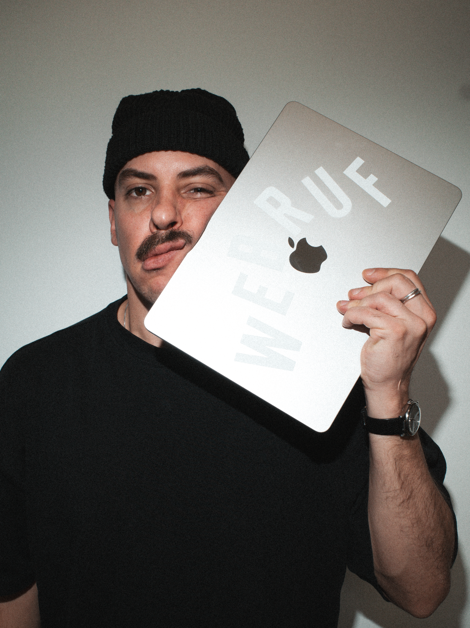Junge Mann trägt schwarze Mütze, trägt schwarzes T-Shirt und trägt eine Uhr, hält ein silbernes Apple MacBook, auf dem Wort "WARRUE" und der Apple-Logo zu sehen sind, vor seinem Gesicht und macht ein Winken mit der linken Hand.