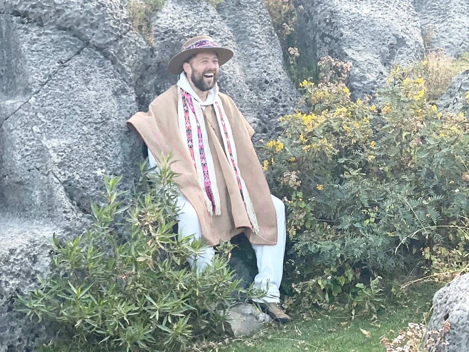 A man sitting on a rock outdoors, smiling, wearing a wide-brimmed hat, a tan cloak with colorful embroidery, white pants, and hiking shoes, surrounded by rocks and bushes.