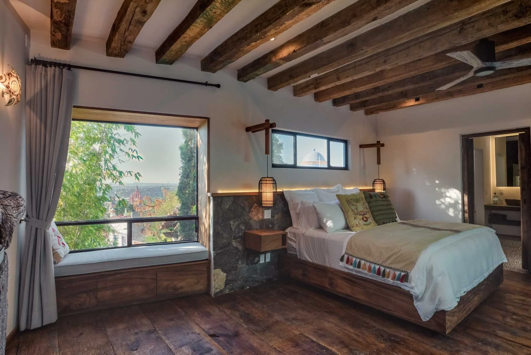 A cozy bedroom with wooden beams on the ceiling, a large bed with white bedding and colorful pillows, a window seat with a pillow, and views of lush greenery outside. There are two hanging lamps on either side of the bed and a small bedside table.