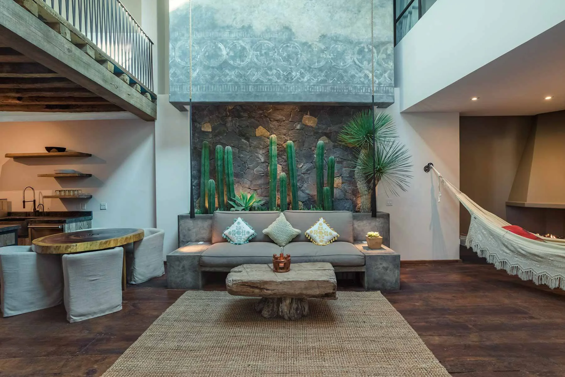 Interior of a modern house with a living room and a second-floor balcony. The room features a stone wall with tall green cacti and a small palm, a beige sofa with decorative pillows, a rustic wooden coffee table, a hammock on the right, and a circula